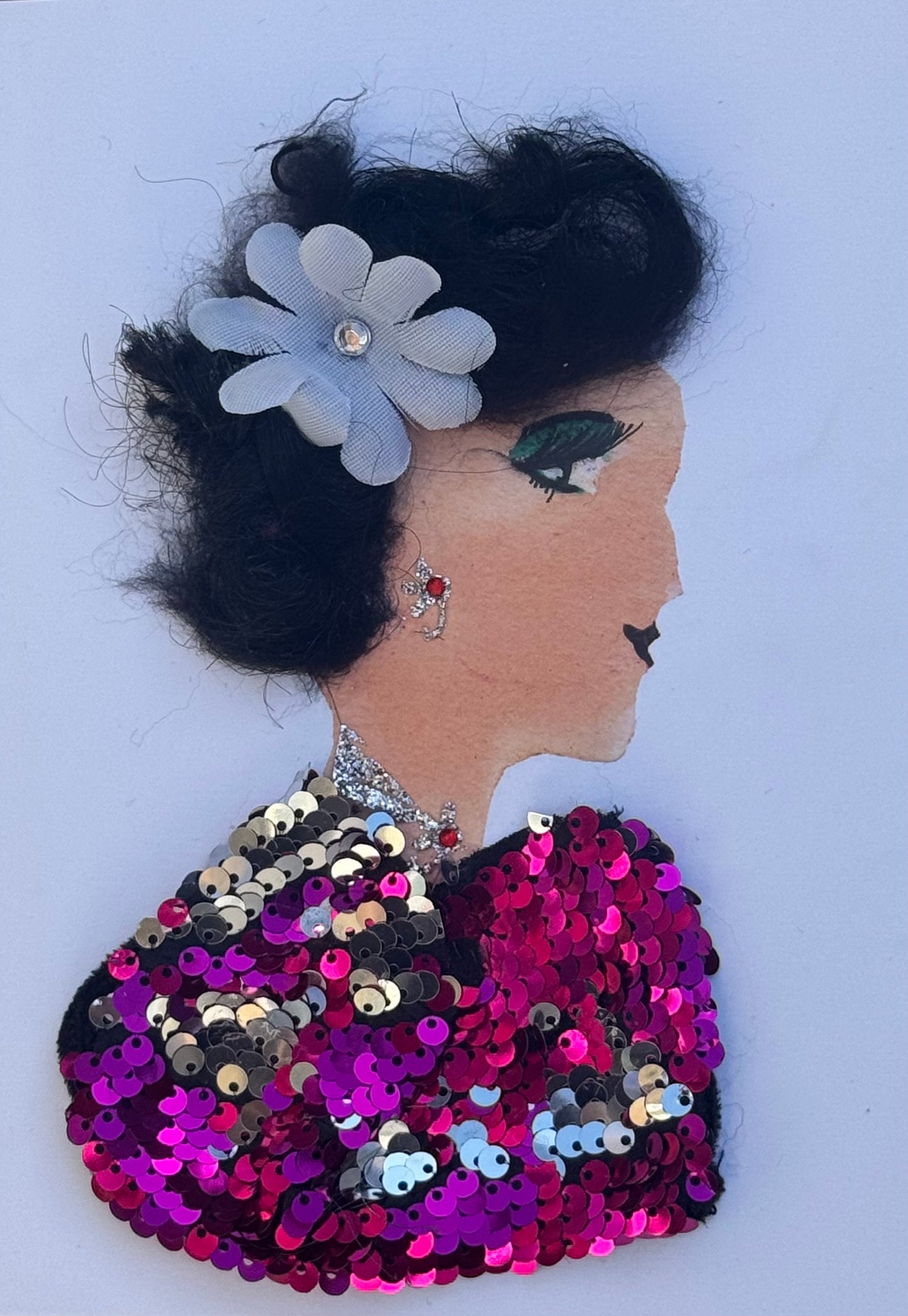 Handmade greeting card that can double as a piece of art featuring a profile collage of a woman with softly curled dark hair styled up and adorned with a white layered flower with a rhinestone center. She wears green eye makeup, delicate earrings, and a shimmering pink, magenta, and silver sequin top that creates a textured, reflective surface. The sequins overlap densely across the bodice, catching light and giving the garment a dimensional, celebratory appearance against the clean white background.