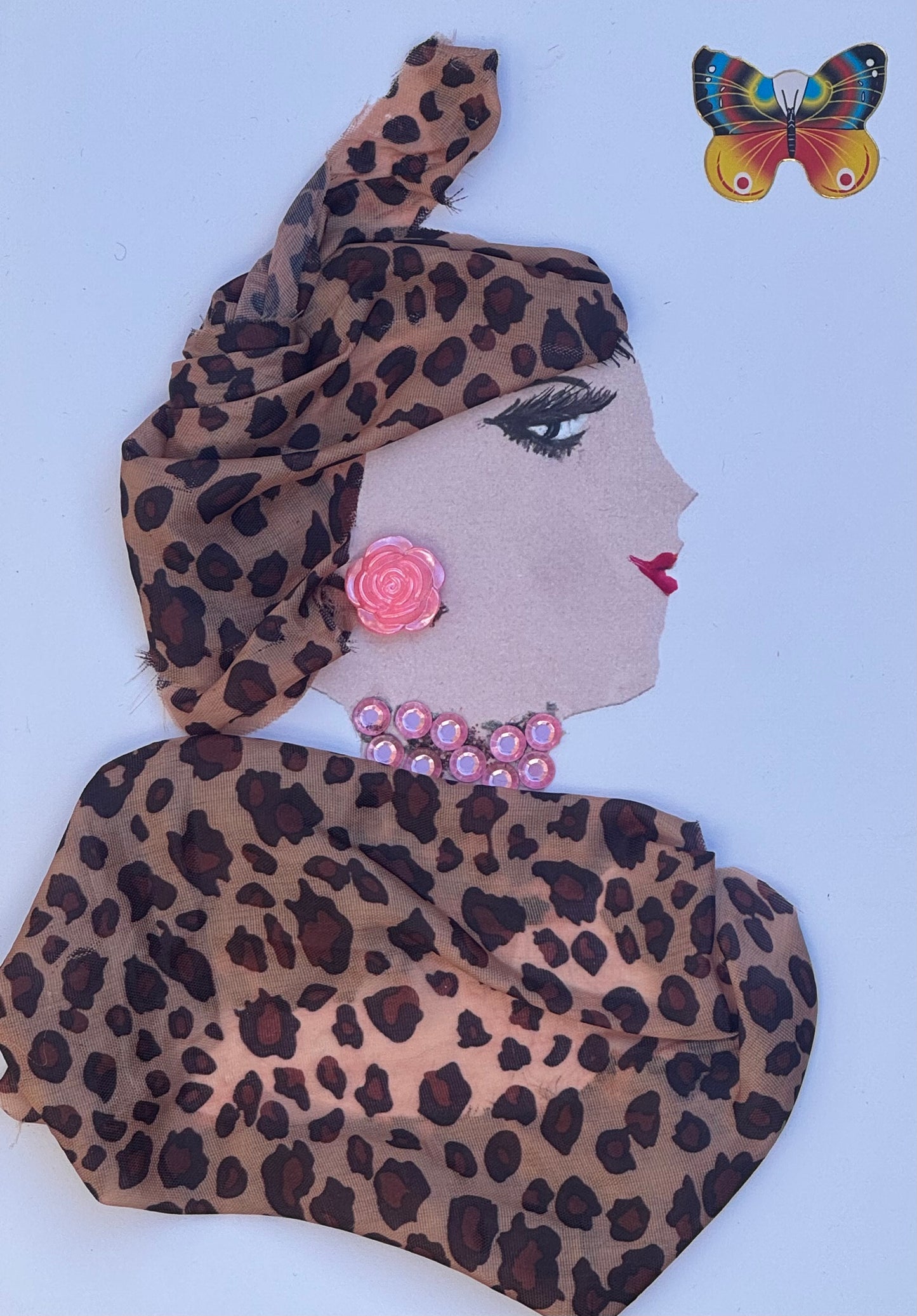 Handmade greeting card that can double as a piece of art featuring a profile collage of a woman wearing a structured leopard print headwrap tied at the top and a matching draped garment. She has a soft pink rose-shaped earring, a pink gem necklace, and defined eye makeup with long lashes and red lips. A small butterfly sticker appears near the top corner, and the layered fabrics create depth and dimension, with gentle folds and shadows giving the piece a sculptural, handcrafted appearance.