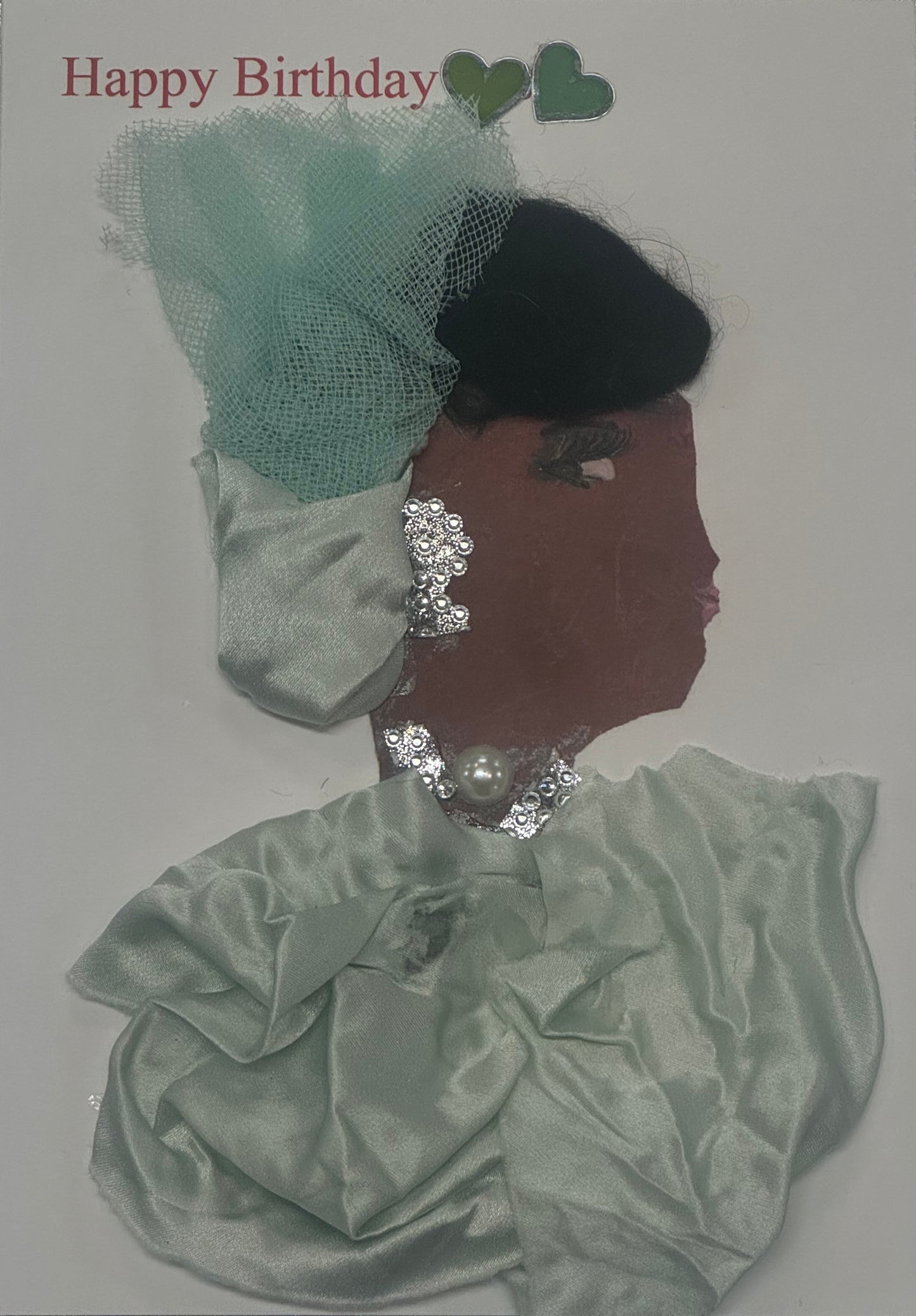 Handmade birthday card that can double as a piece of art, featuring a woman wearing a mint green satin outfit with a decorative tulle fascinator hat. She is adorned with pearls and sparkling silver jewelry, with the neckalce and earrings matching, giving the design a soft pastel elegance. Above the ladys head there are also two green colored hearts that sit in the top middle, next to the written "Happy Birthday" in the top left of card.