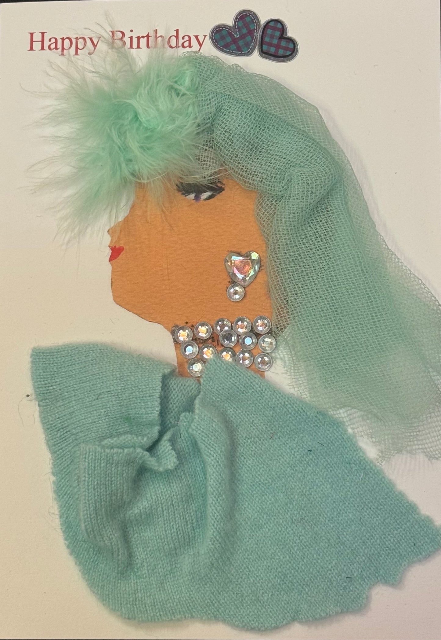 Handmade greeting card that can double as a piece of art featuring a side-profile of a woman wearing a decadent mint green textured head scarf with a feather detail and also wearing a matching green shawl made up of sweater material. She also has rhinestone jewelry on, with a bedazzled necklace and heart shaped earrings. There is a “Happy Birthday” text with purple and mint green heart decorations above her.