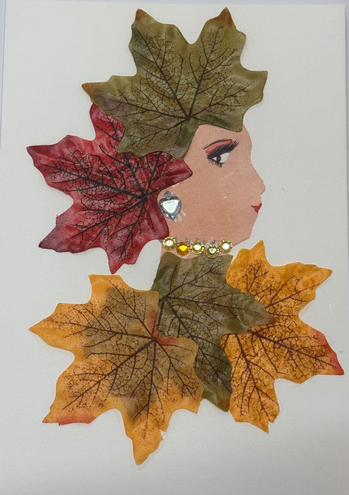 Handmade greeting card that doubles as a piece of art showing a woman in profile, with her hair formed from layered autumn leaves in rich shades of red, orange, and gold. The leaves overlap and fan outward, creating a textured, flowing silhouette that mimics natural hair movement. She wears a coordinating outfit that blends with the autumn palettee. She also wears a a gold gem neckalce and a silver heart shaped gem as earrings, contrasting with the matte leaf textures.