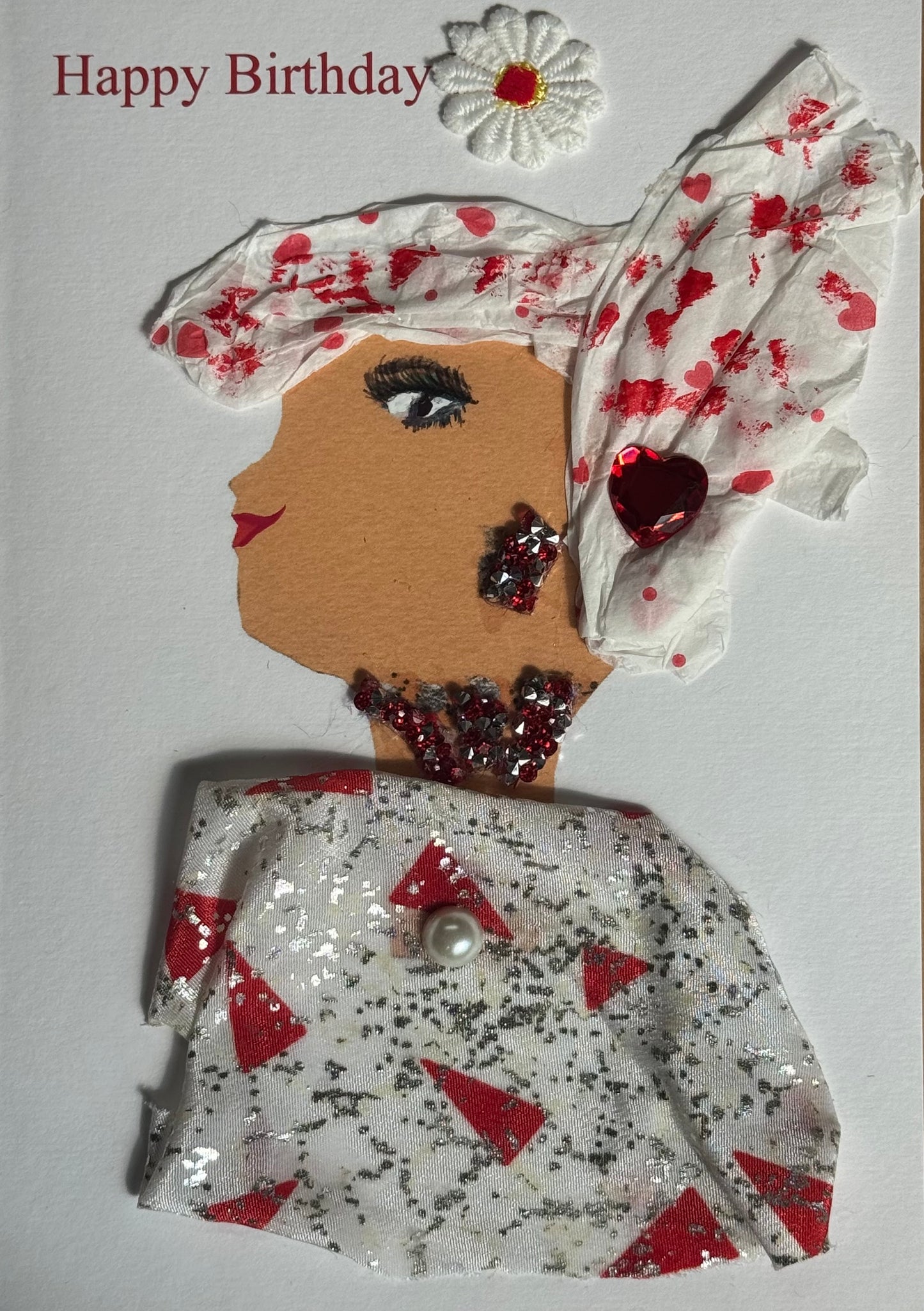 A handmade birthday card showing a woman in profile with medium brown skin facing right. She wears a white headwrap with red splatter-style accents and a white flower embellishment above. Her jewelry includes a red heart gem earring and a red beaded necklace. Her top is made from white fabric covered with silver glitter and red triangular shapes. A white flower sticker appears above her head and “Happy Birthday” is printed in red at the top on a white background.