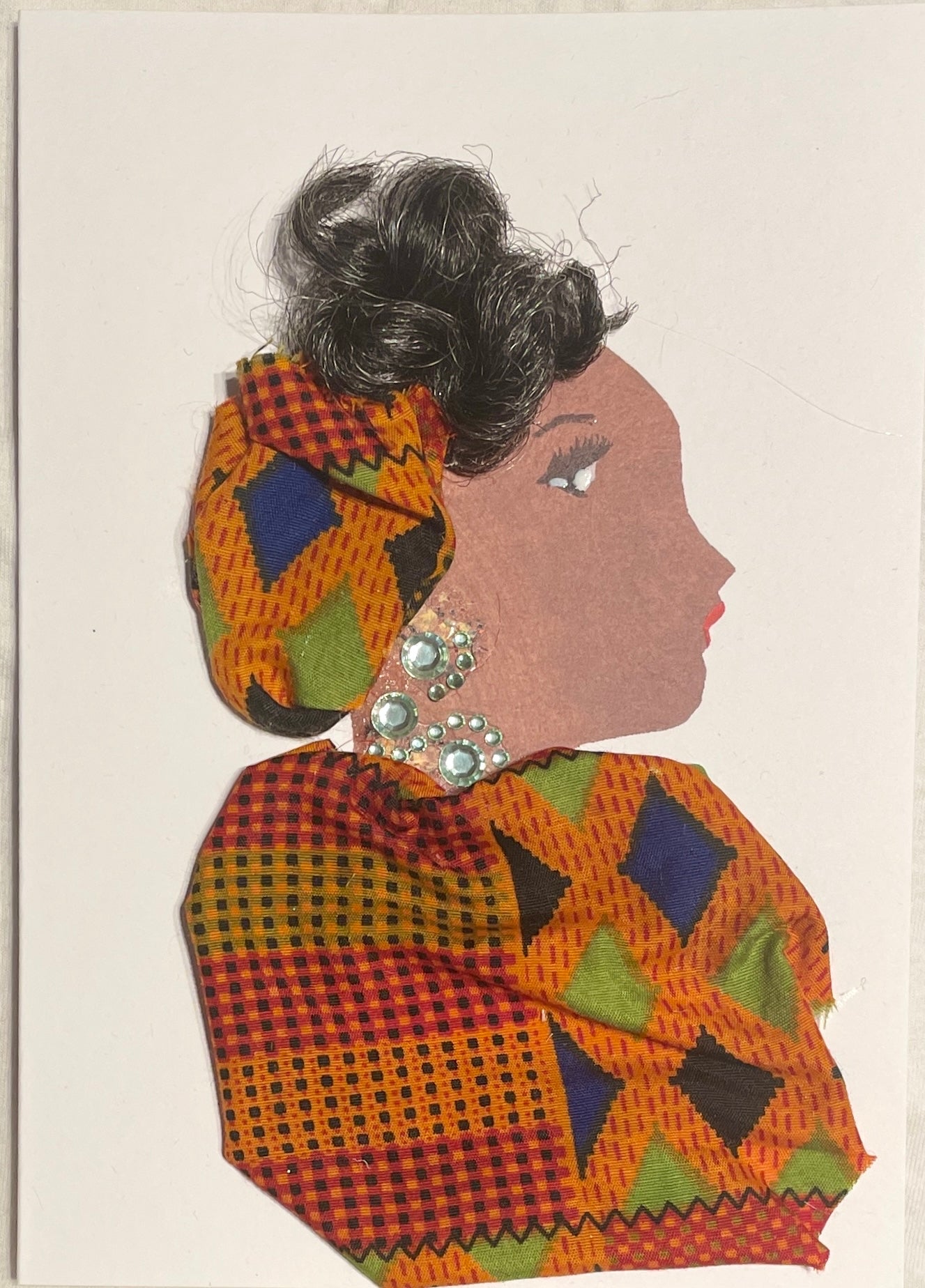 This is a handmade card of a woman named Aduke American. Her hair is styled in a swept-up bun, wrapped partly in colourful fabric that matches her outfit. She wears sparkling gem earrings and a matching necklace. Her outfit is made from bold patterned fabric in red, orange, blue, green, and black, featuring geometric shapes and fine detailing.