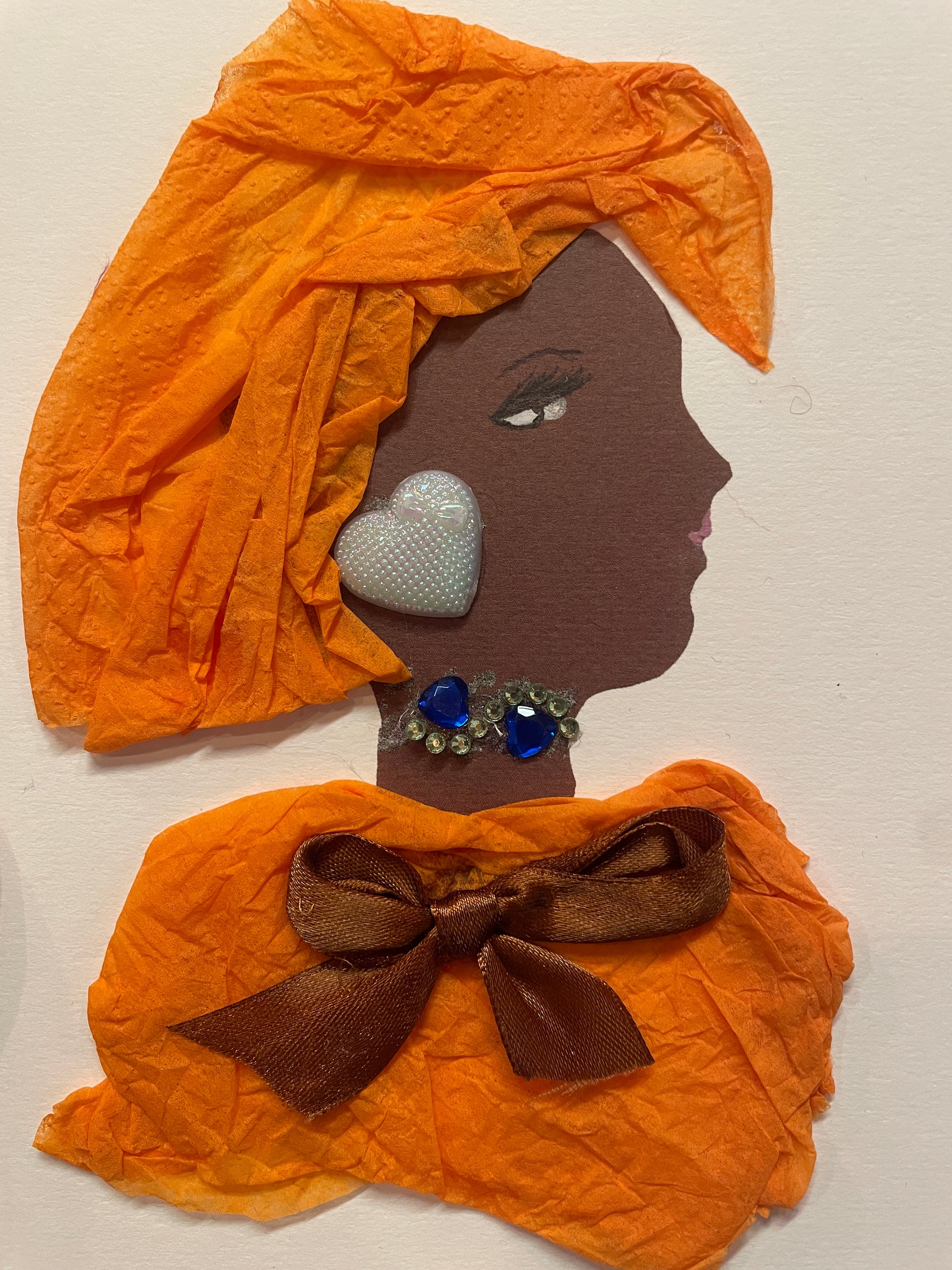 A  handmade card of a woman in profile wearing textured orange tissue as a head wrap and top. She has a brown bow on her chest, a large pearl heart-shaped earring, and a blue jewelled necklace.