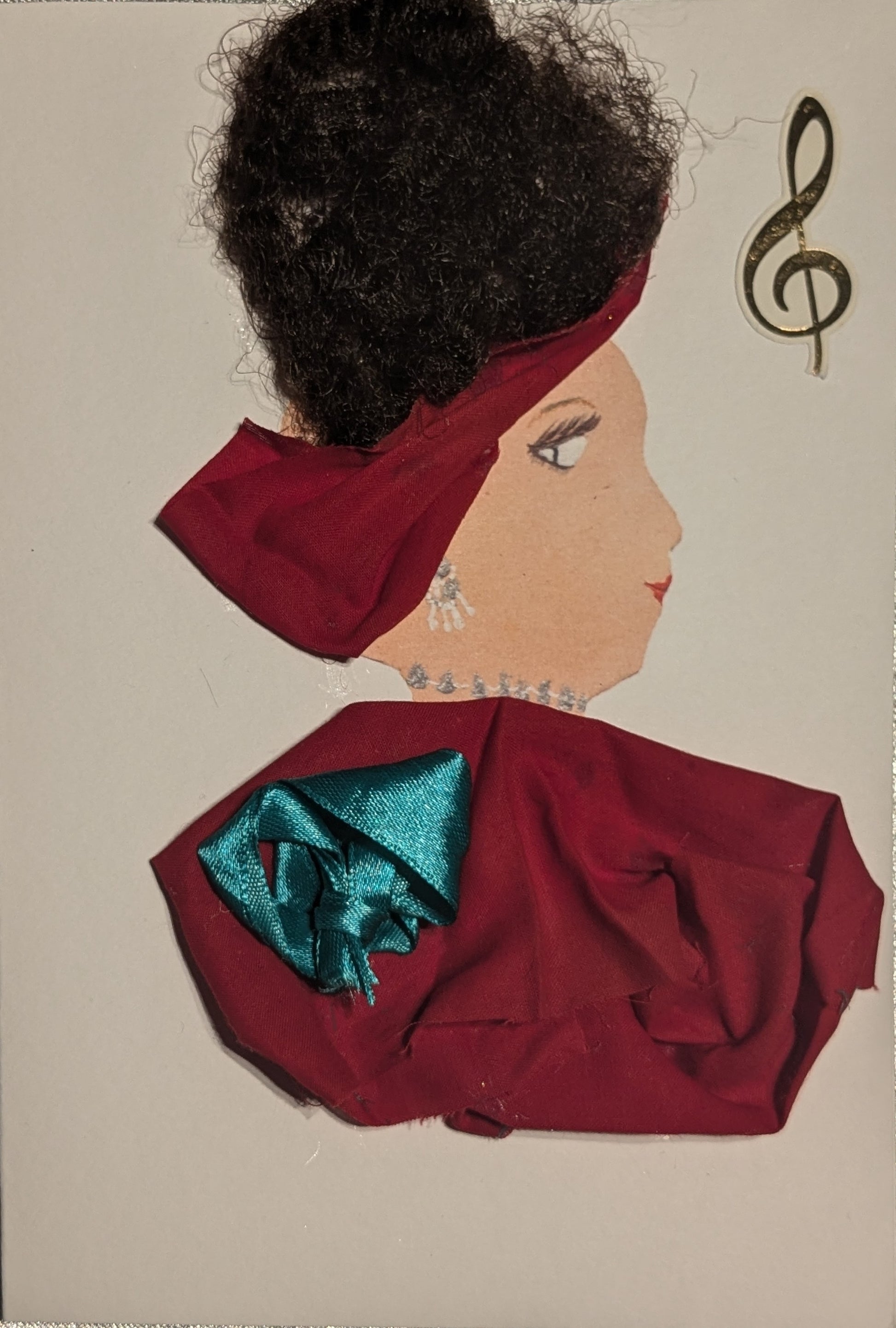 I handmade this card of a woman named Amiee Amaranthus. She’s shown in profile with expressive eyes, red lips, and voluminous black textured hair wrapped with deep red fabric. She wears sparkling silver accents as earrings and along her neckline. Her outfit is fashioned from the same red fabric, adorned with a bold teal satin rosette. A golden treble clef sticker floats beside her, hinting at her musical spirit.