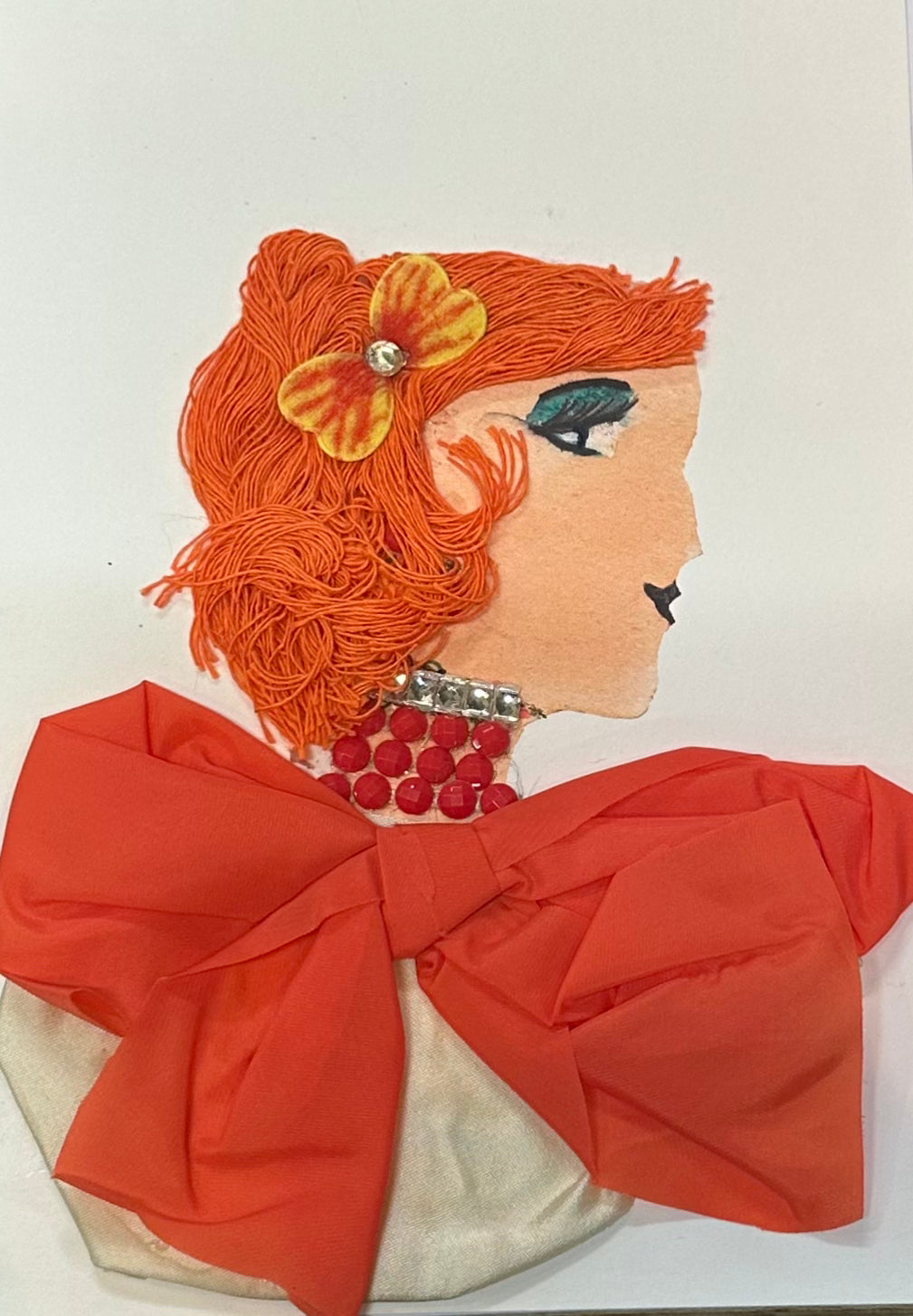 A handcrafted portrait of “Orange Aunty,” featuring bright orange yarn hair with a yellow flower clip, bold green eye makeup, a red beaded necklace, and a large orange fabric bow forming the outfit.