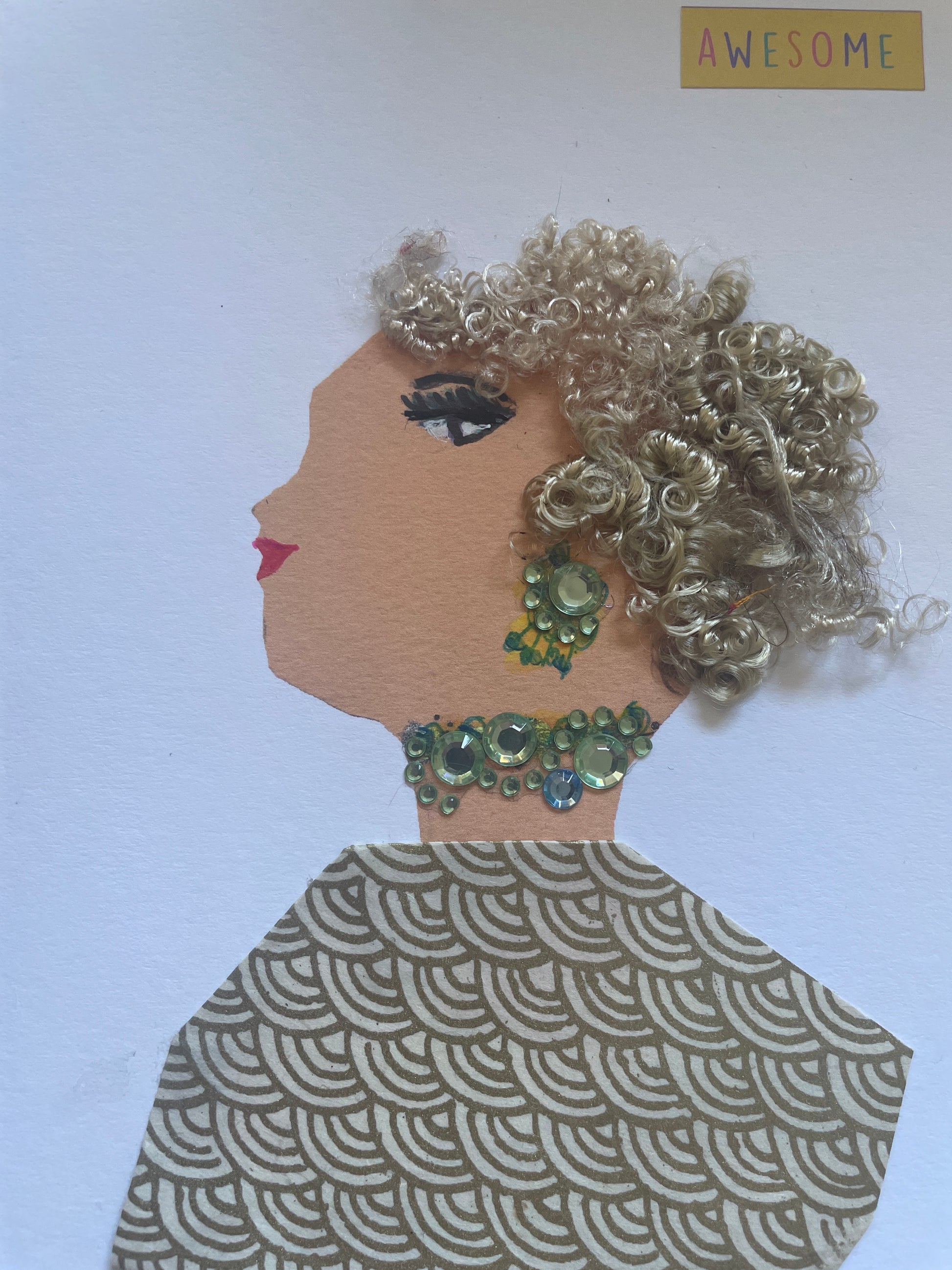 I handmade this card of a woman named Awesome Lemonade. She’s shown in profile with shimmering silver curls, dramatic lashes, and soft red lips. Her earring and necklace are made of pale green rhinestones, adding an elegant sparkle. She wears a gold and cream geometric patterned garment. A small “AWESOME” sticker floats above her, perfectly matching her confident, graceful energy.