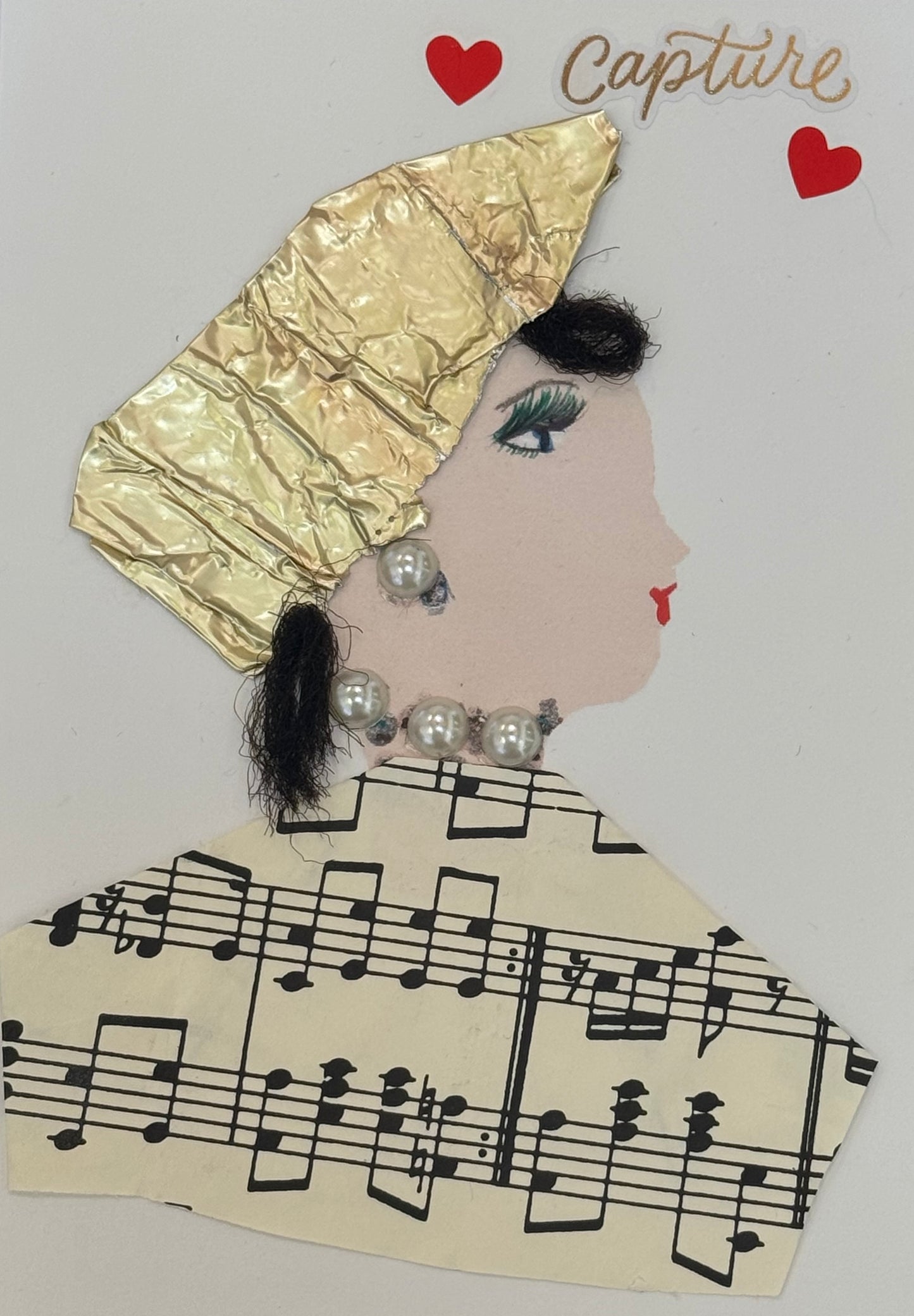 Handmade greeting card that can double as a piece of art. Featuring a side profile of a fair skinned woman. She wears a ttextured metallic gold headwrap that covers the top of her head. There are three stickers: a heart and a sticker with capture and another heart. she has short black curled hair , and bright red lips. She is accessorised with large pearl earrings and a matching pearl necklace composed of oversized beads.She has a musical top