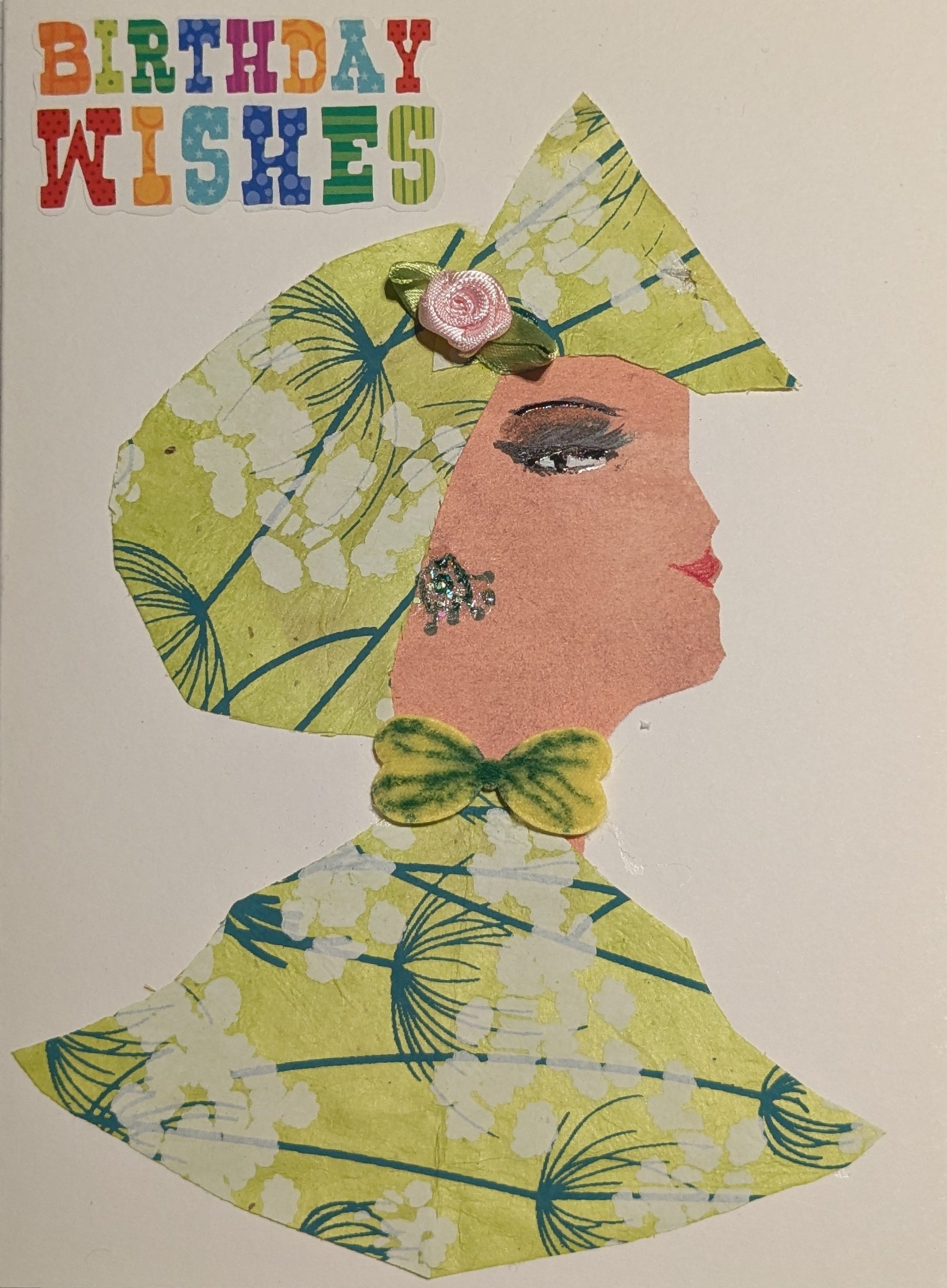 This is a handmade card of a woman named Belladonna Baby's Breath. She wears a matching hat and outfit made from light green paper with a floral pattern of white blossoms and blue stems. A small pink rose embellishes her hat, and a green bow is tied at her neck. Her earring is made from shiny gems, and her face is drawn in profile with defined eyes and red lips. At the top left corner, colourful letters spell out “Birthday Wishes.”