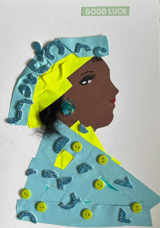 Here is Blakeley Blue, a striking vision of creativity and sparkle. Her outfit and headwrap are pieced together from soft powder-blue paper, with bold flashes of neon yellow that echo through her collar and turban. Scattered across her look are glittery turquoise shapes and cheerful yellow buttons—each one adding playful texture. A shimmering blue earring and a calm “GOOD LUCK” message above her complete the composition