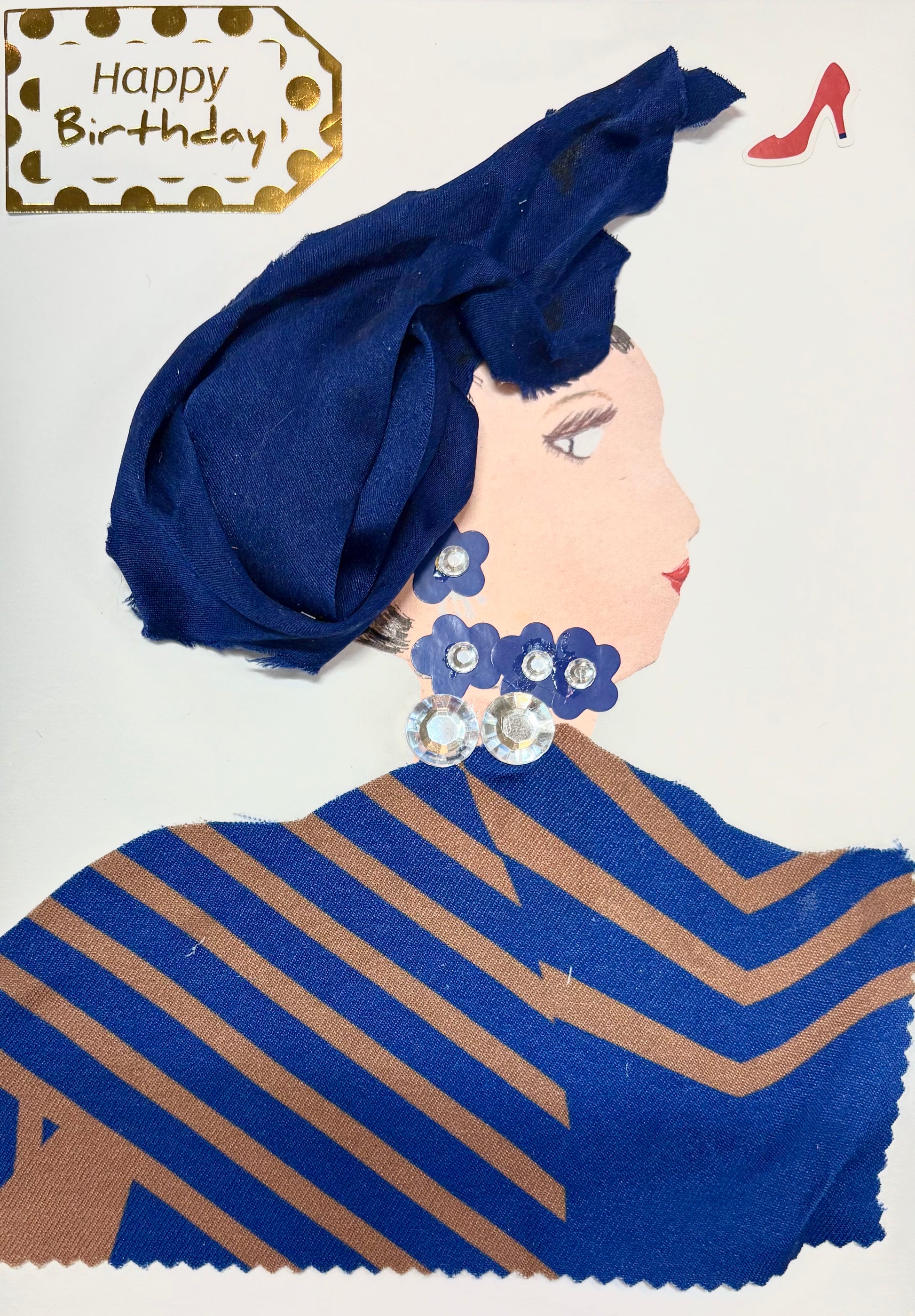 This is a handmade card of a woman named Blue Bethany. She wears a deep blue headwrap and a matching outfit made from fabric with tan diagonal stripes. Her jewellery includes sparkling gems arranged with navy blue flower shapes for her earrings and necklace. A “Happy Birthday” tag with gold dots appears in the top corner, along with a small red high-heeled shoe sticker.