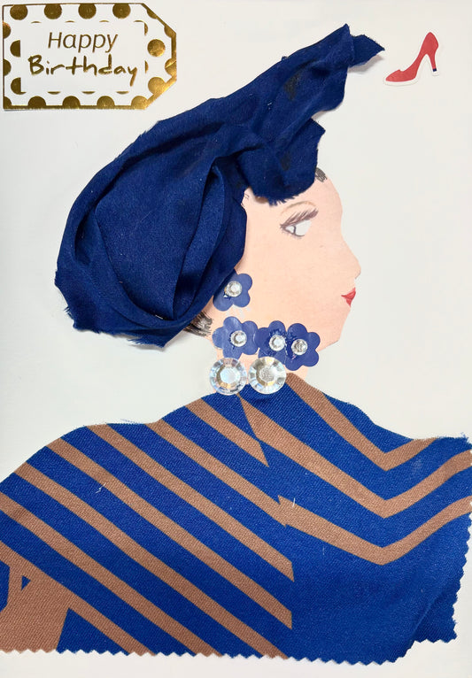 This is a handmade card of a woman named Blue Bethany. She wears a deep blue headwrap and a matching outfit made from fabric with tan diagonal stripes. Her jewellery includes sparkling gems arranged with navy blue flower shapes for her earrings and necklace. A “Happy Birthday” tag with gold dots appears in the top corner, along with a small red high-heeled shoe sticker.