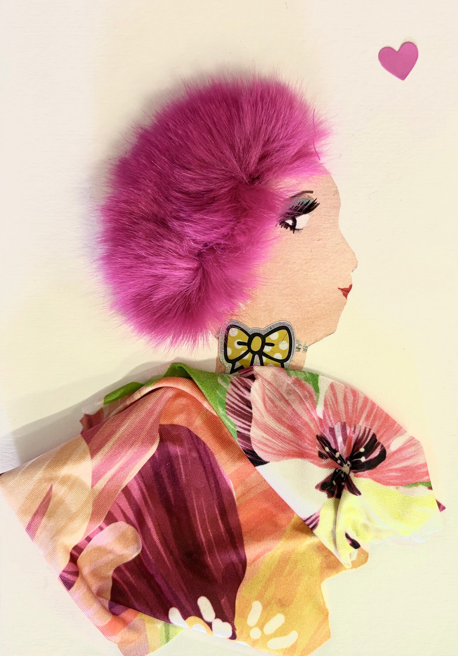 Handmade portrait of a woman in profile. She has bright pink fluffy hair, red lipstick, and wears a yellow bow earring. Her outfit is made from colourful floral fabric, and there is a small pink heart shape near the top right corner.