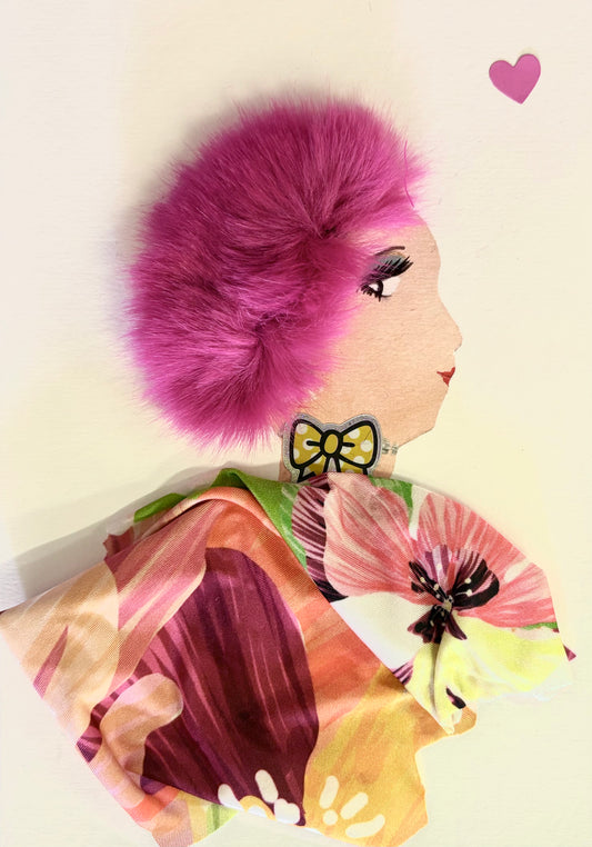 Handmade portrait of a woman in profile. She has bright pink fluffy hair, red lipstick, and wears a yellow bow earring. Her outfit is made from colourful floral fabric, and there is a small pink heart shape near the top right corner.