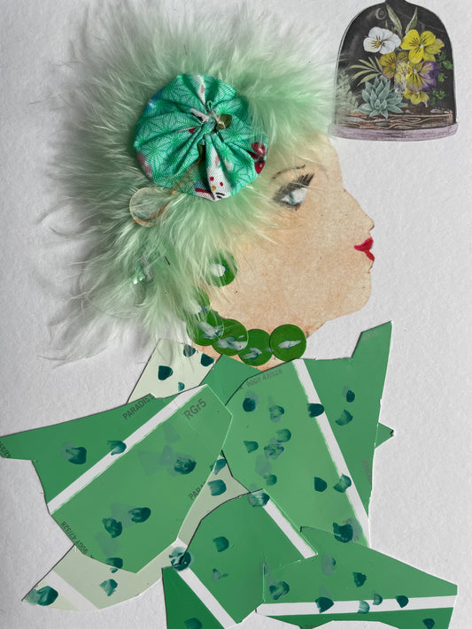 I handmade this card of a woman named Greenwich Flora. She’s shown in profile with soft makeup, red lips, and a dreamy gaze. Her hair is made of pale green feathers, accented with a gathered green floral fabric and a translucent bead. She wears a necklace of green discs and an outfit crafted from green paint swatches, hand-dotted with teal paint. A sticker of a glass-domed flower arrangement floats nearby.
