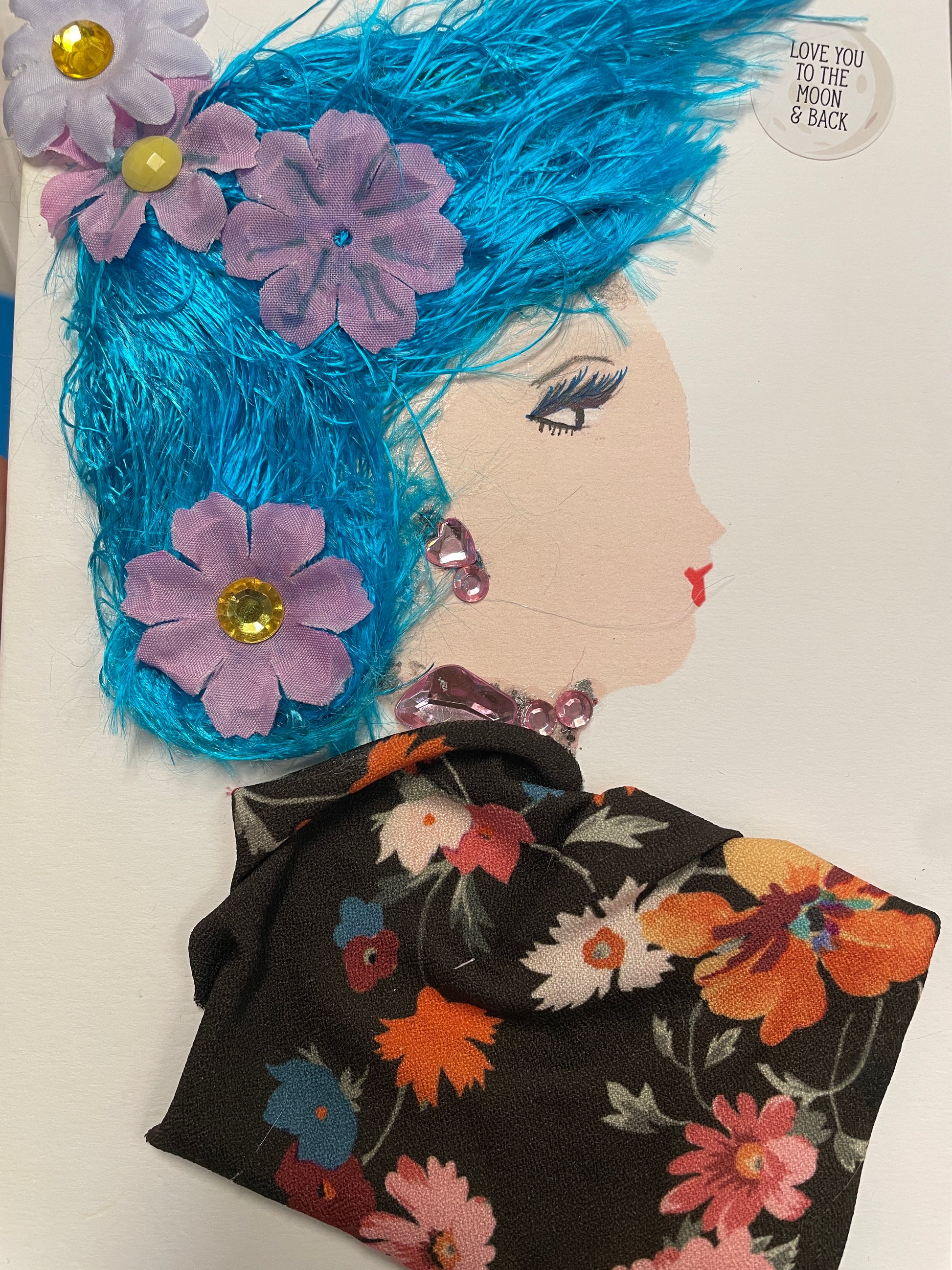 Handmade card showing a woman in profile with bright blue textured hair, decorated with purple fabric flowers and gem centers. She wears pink crystal earrings and a necklace, and a black floral dress with red, blue, and orange flowers. A circular sticker near her head reads, “Love you to the moon & back.”