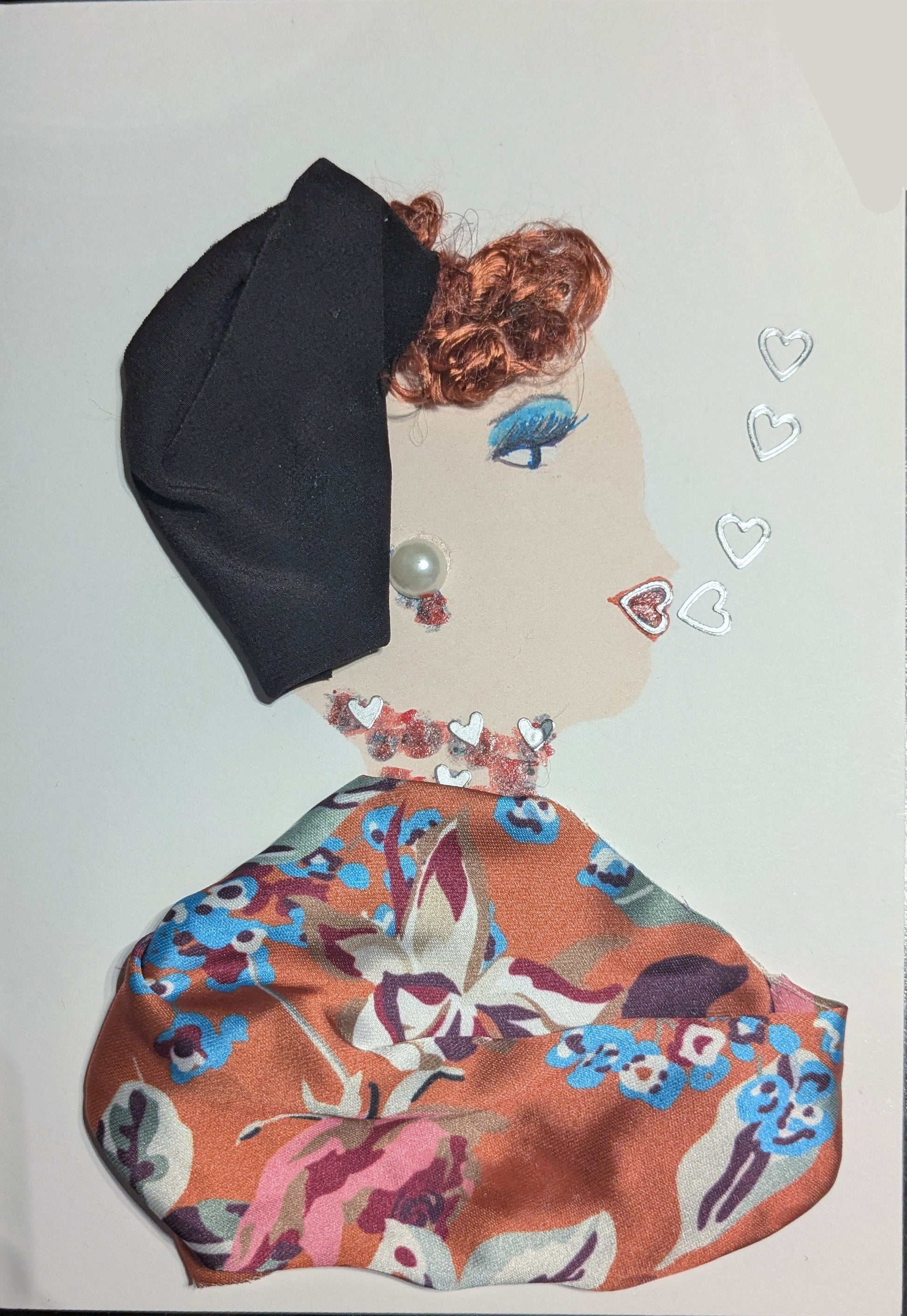 This is a handmade card of a woman named Camden Chapter. She has curly auburn hair partially covered by a folded black headwrap. A pearl adorns her ear as an earring, and she wears a necklace made of red and white heart-shaped accents. Her outfit is made from silky fabric with a bold floral pattern in orange, blue, pink, and white. A few small outlined hearts float beside her face.