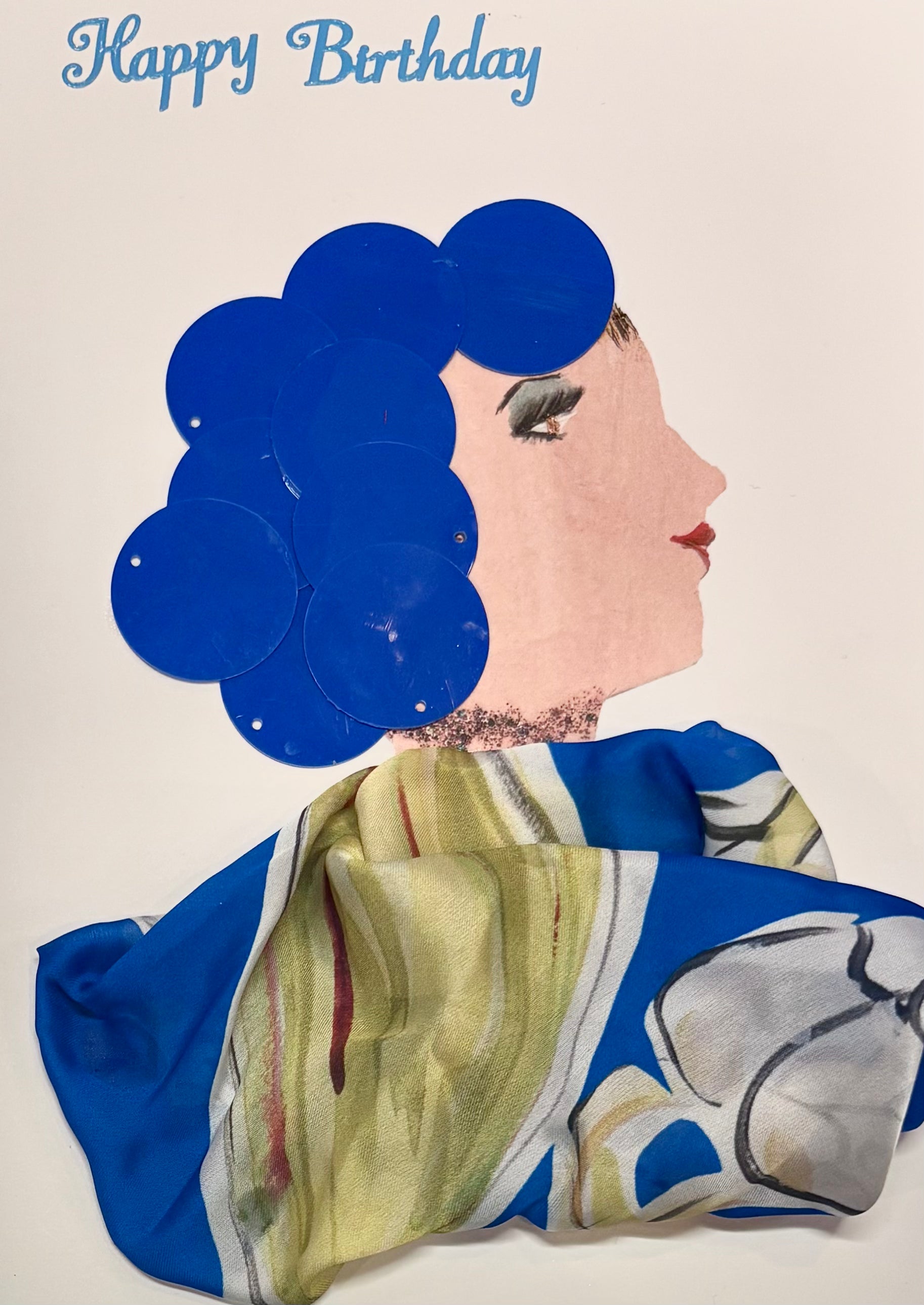 This is a handmade card of a woman named Cerulean Cecilia. Her hair is styled using large, shiny blue sequins arranged in soft waves. She wears a flowing fabric wrap in shades of blue, green, white, and hints of red, featuring an abstract floral print. At the top of the card, the words “Happy Birthday” are written in light blue script.