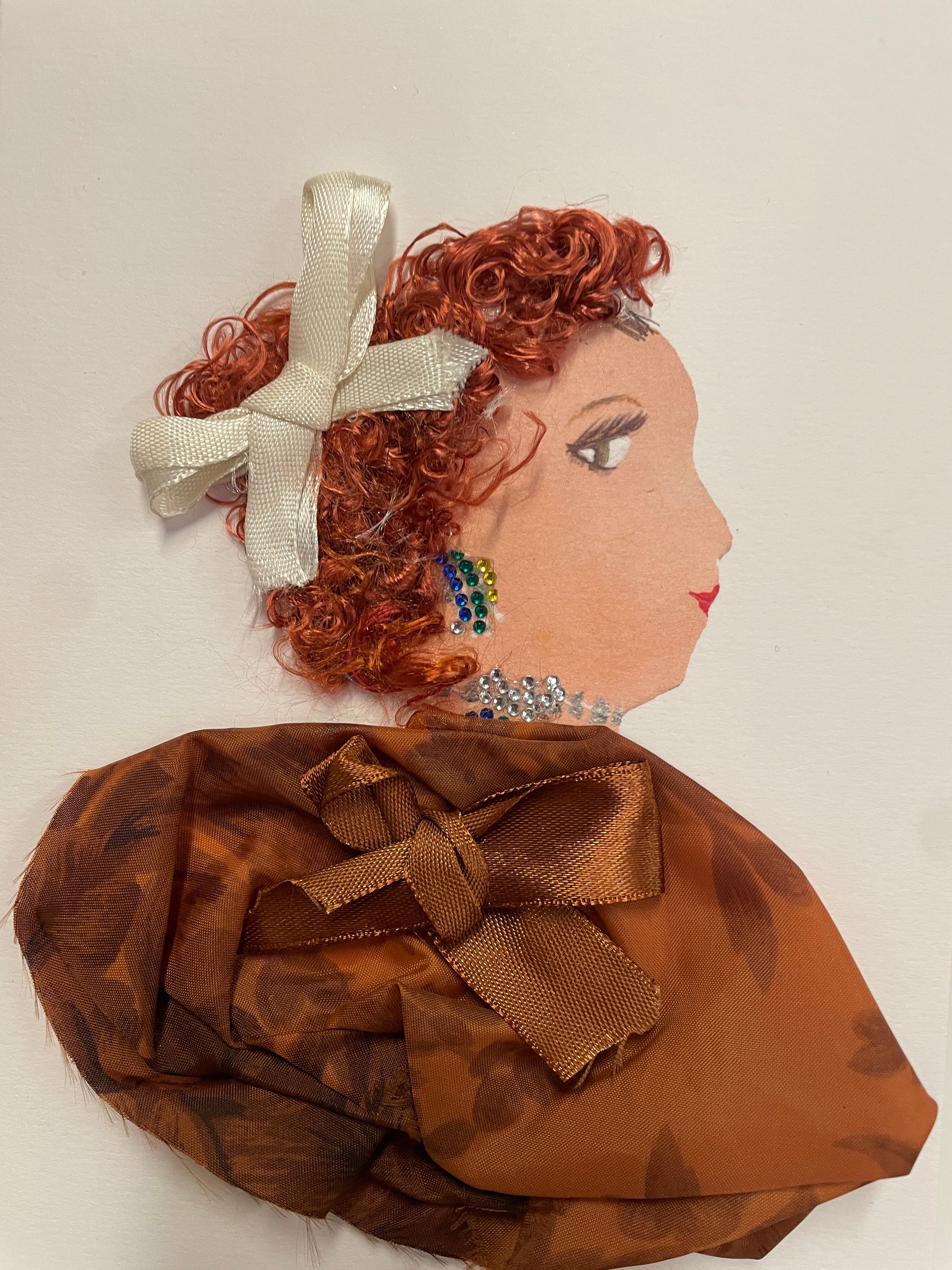 Handmade greeting card featuring a side-profile of a woman with curly red hair adorned with a cream ribbon bow. She wears colorful beaded earrings, a sparkly silver necklace, and a rich chestnut brown fabric top with a large satin bow. The card has a textured, dimensional design with a white background.