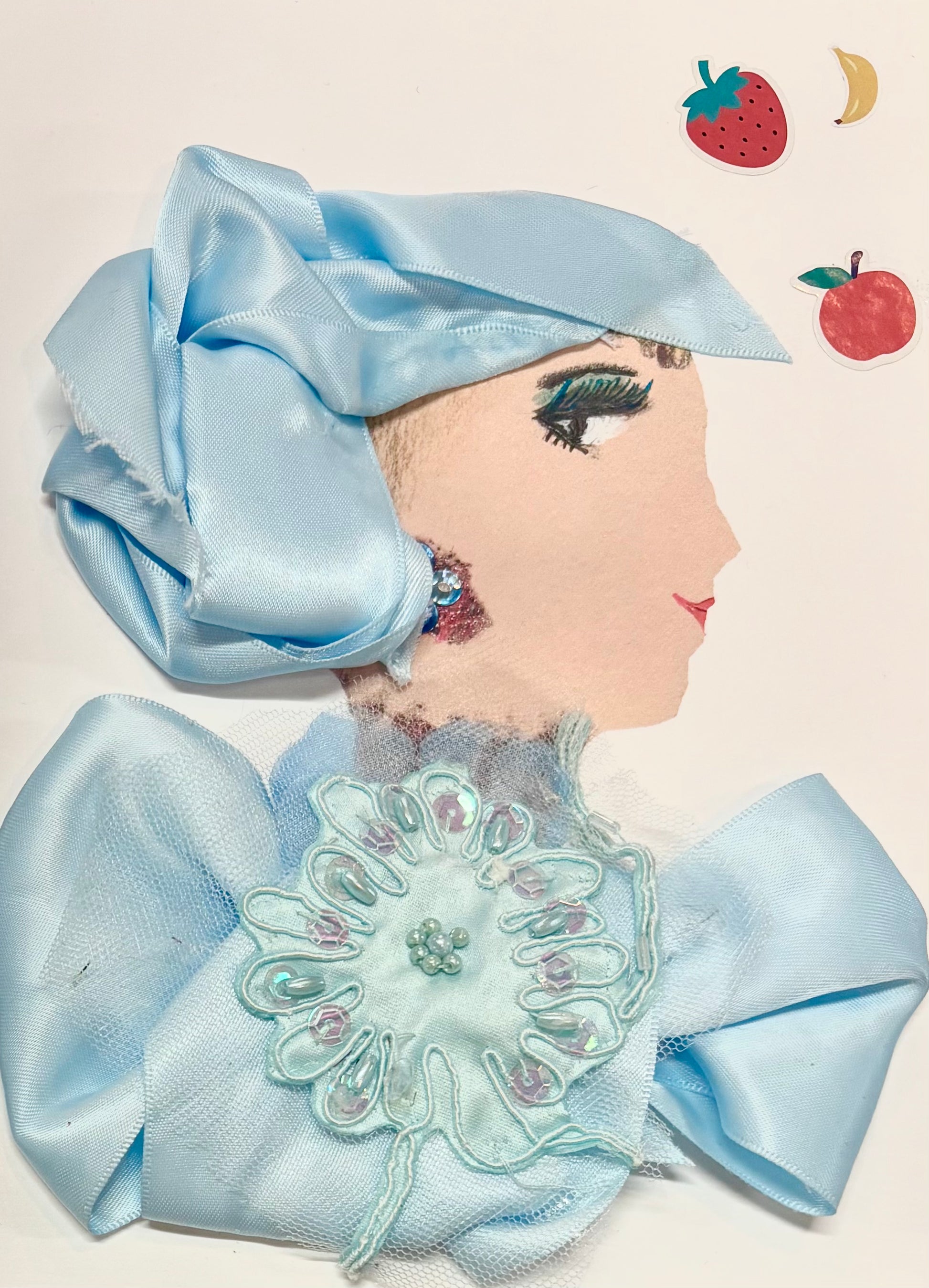 A woman wears a light blue silk headwrap and a matching blouse. Her blouse has a large light blue flower in the center. She wears a subtle blue earring. In the top right corner, there is a strawberry, banana, and apple.