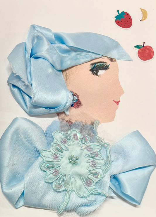 A woman wears a light blue silk headwrap and a matching blouse. Her blouse has a large light blue flower in the center. She wears a subtle blue earring. In the top right corner, there is a strawberry, banana, and apple.