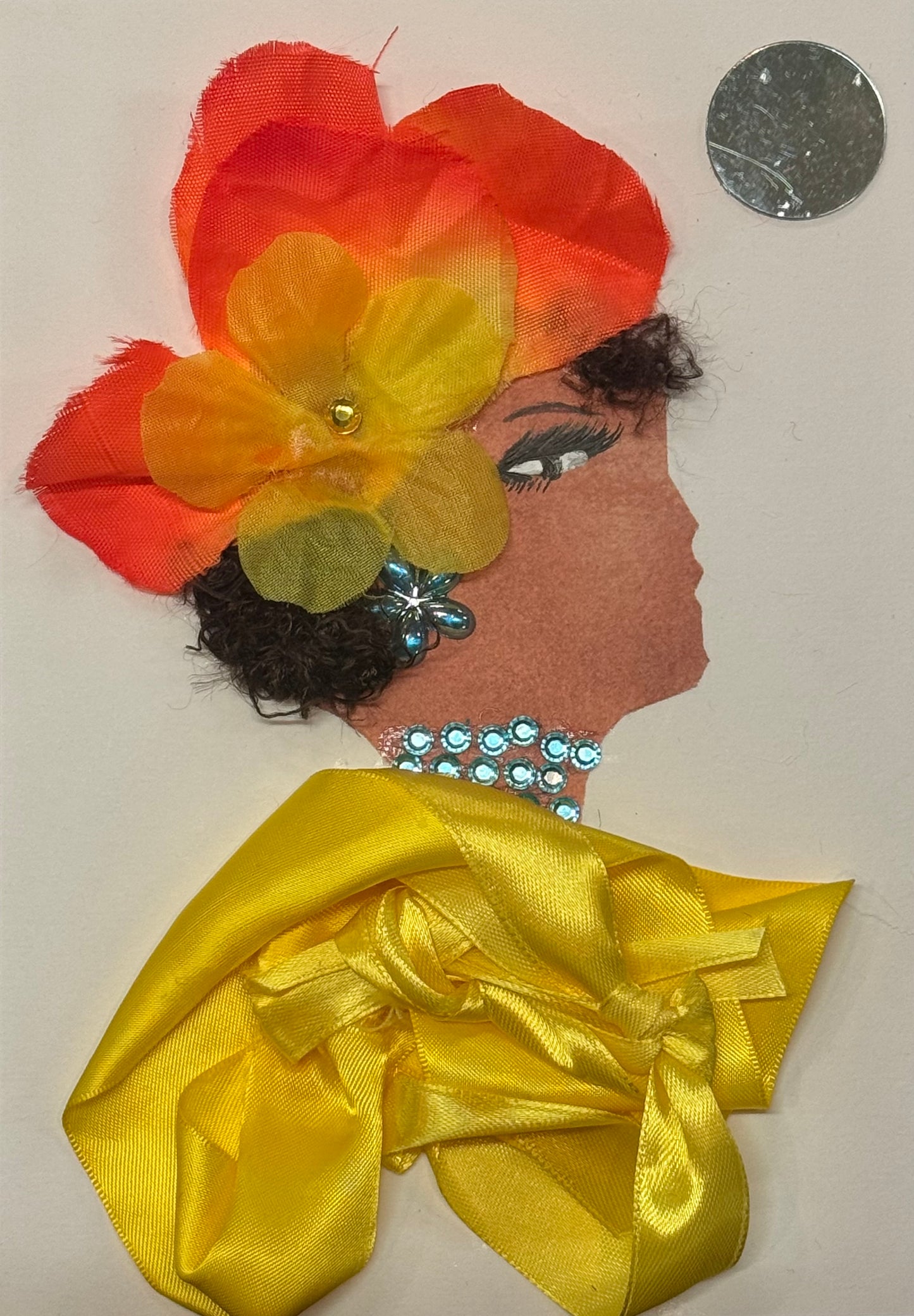 Handmade greeting card that can double as a piece of art, featuring a side-profile silhouette with curly dark hair, adorned with a large layered orange and yellow fabric flower that is ombre coloured with orange and yellow and has a small yellow gem in the centre. She also wears blue gem earrings and necklace. For her shawll she wears a bright yellow satin top that has a bow garment in the middle. Above her head on the right, the is a circular reflective gem that acts as a mini mirror.