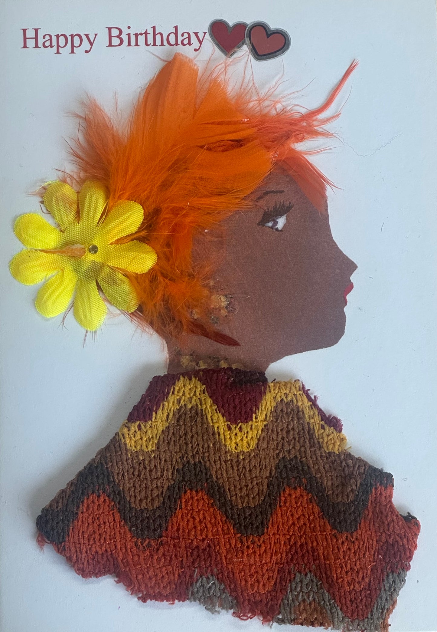 A handmade birthday card featuring a side-profile illustration of a woman wearing an orange feather headpiece with a large yellow flower. Her clothing is made from textured knit fabric in brown and orange zigzag patterns. She has gold-toned gem earrings and red lips. The background is white with “Happy Birthday” printed above and two heart stickers near the top.