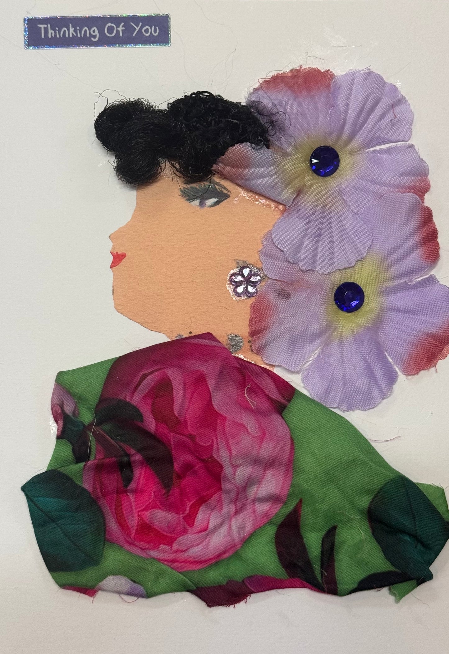 Handmade greeting card that can double as a piece of art, featuring a woman with dark textured hair adorned with large purple flowers. She wears a floral-patterned garment with a prominent rose design and gem accents, with a “Thinking of You” sticker in the top right corner above her head.