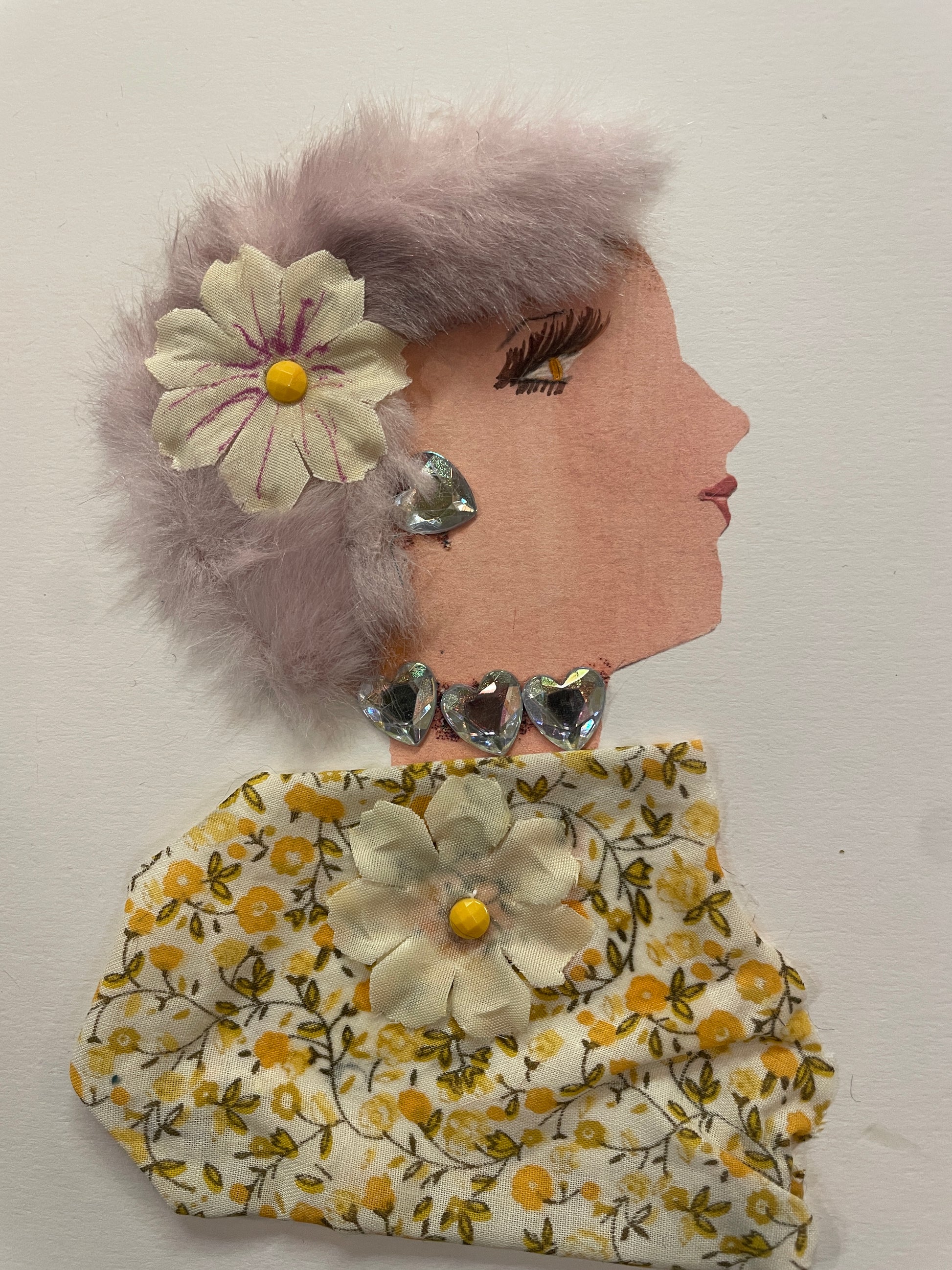 This is a handmade card of a woman named Daisy Dagenham. She has lavender-coloured fuzzy hair, a cream flower with purple lines, heart-shaped gems at her neck, and a yellow floral blouse.