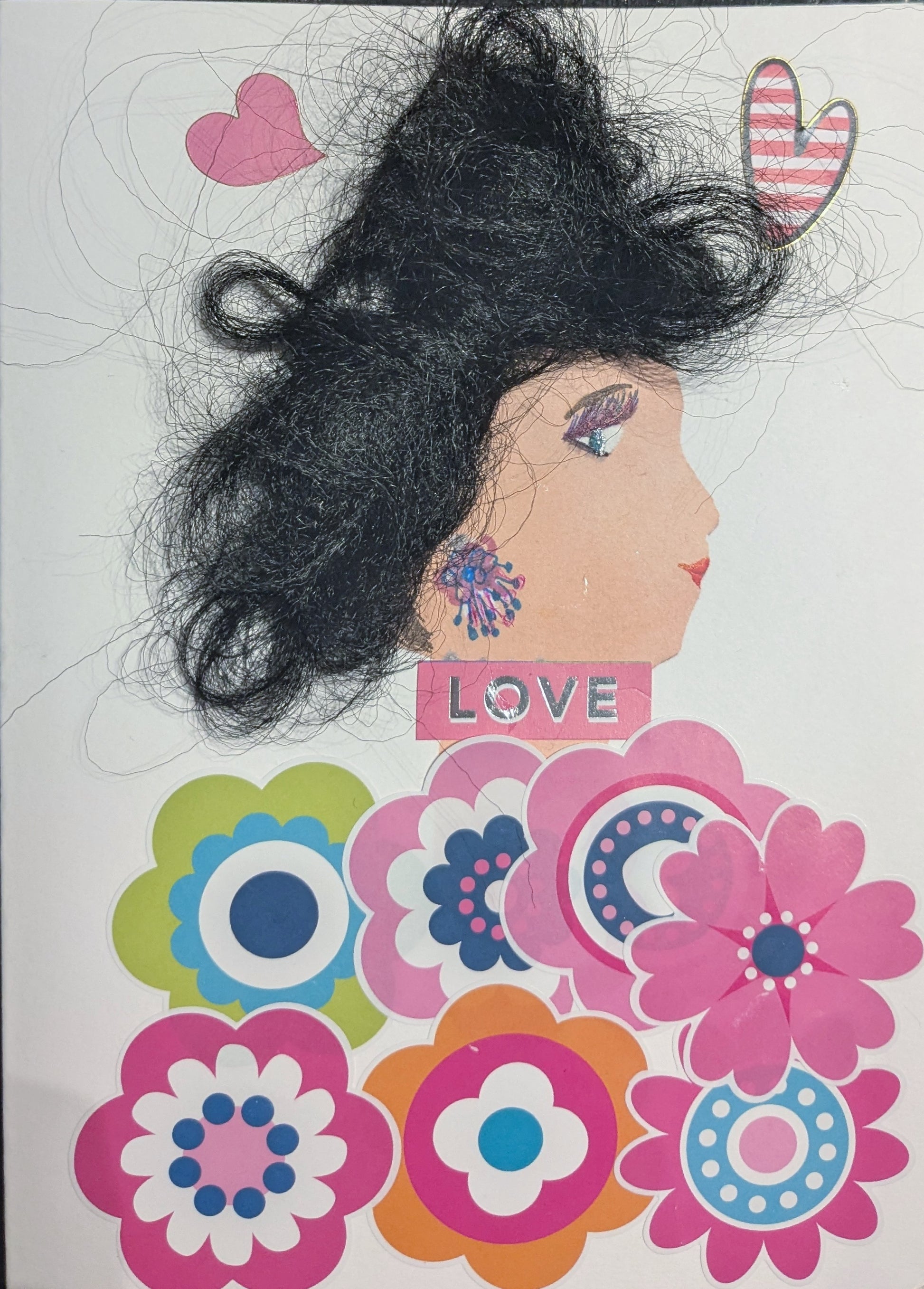 This is a handmade card of a woman named Dalston Daisy. Her hair is made from textured black fibres, styled in a voluminous shape. She wears a sparkling purple and blue earring. Her outfit is composed of brightly coloured flower cutouts in pink, orange, green, blue, and white. Two heart stickers float near her head, and the word “LOVE” is placed in bold letters beneath her earring.