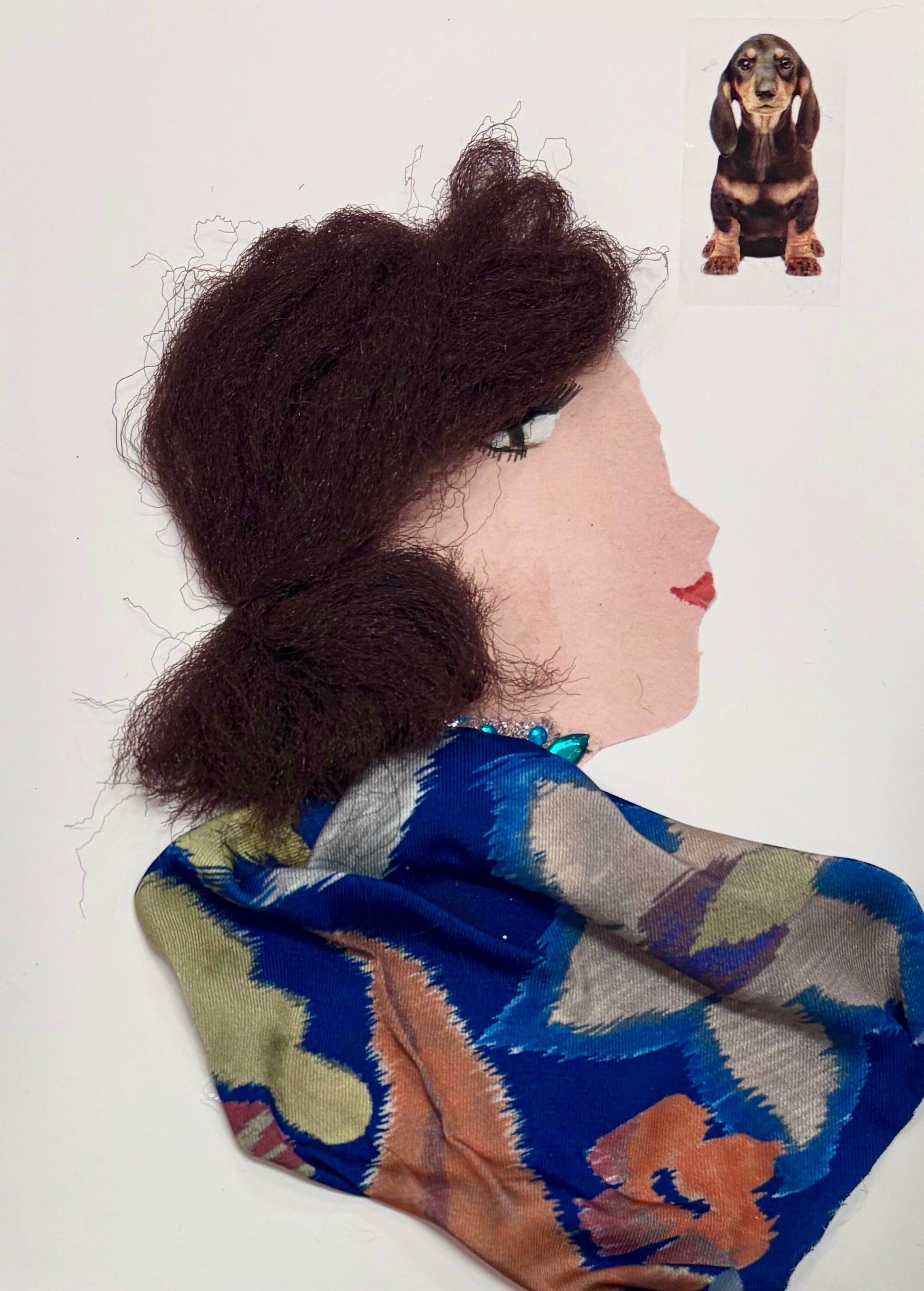 I handmade this card of a woman named Doctor Krista Kilburn. She’s shown in profile with fluffy textured brown hair, soft makeup, and a calm expression. She wears a vibrant blue patterned fabric with floral tones of green, orange, and grey, and a touch of turquoise beading at her neck. A sticker of a dachshund is placed nearby.