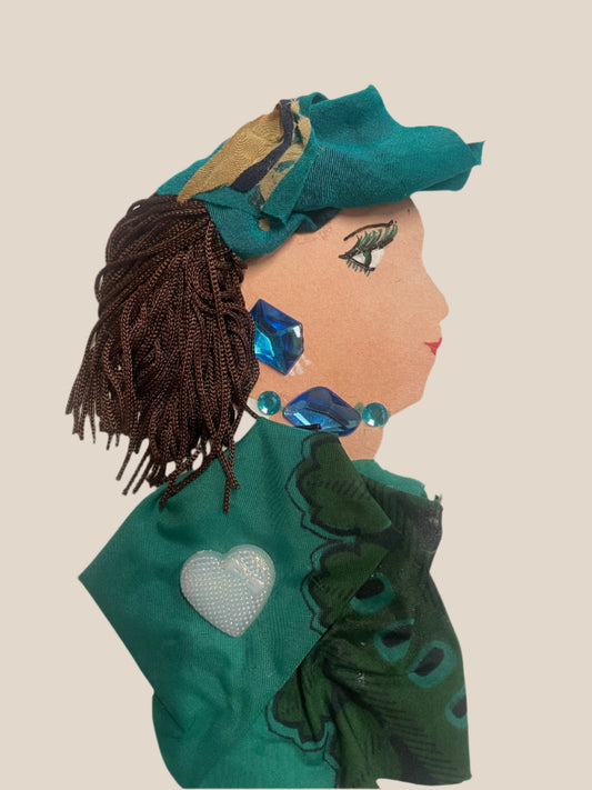 This is a handmade card of a woman named Emerald Eltham. She wears a teal head wrap with brown braided hair, large blue gems for earrings and necklace, and a silky green garment with a white heart embellishment.
