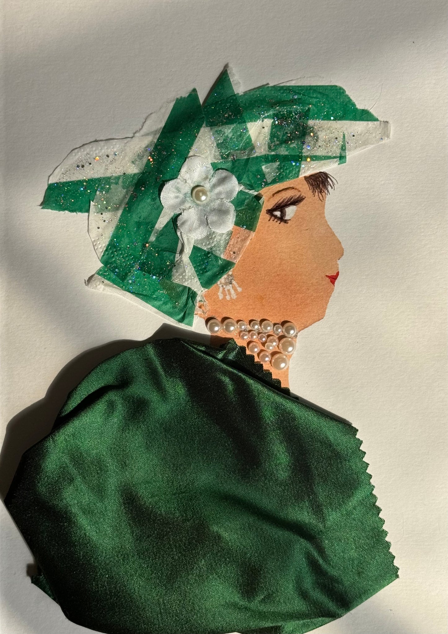 A handmade card showing a woman with medium brown skin in profile facing right. She wears a layered green ribbon hat with white accents and a white flower embellishment. Her jewelry includes pearl earrings and a pearl necklace. Her shoulders are draped in dark green satin fabric. The background is white with “Happy Birthday” printed in red at the top.