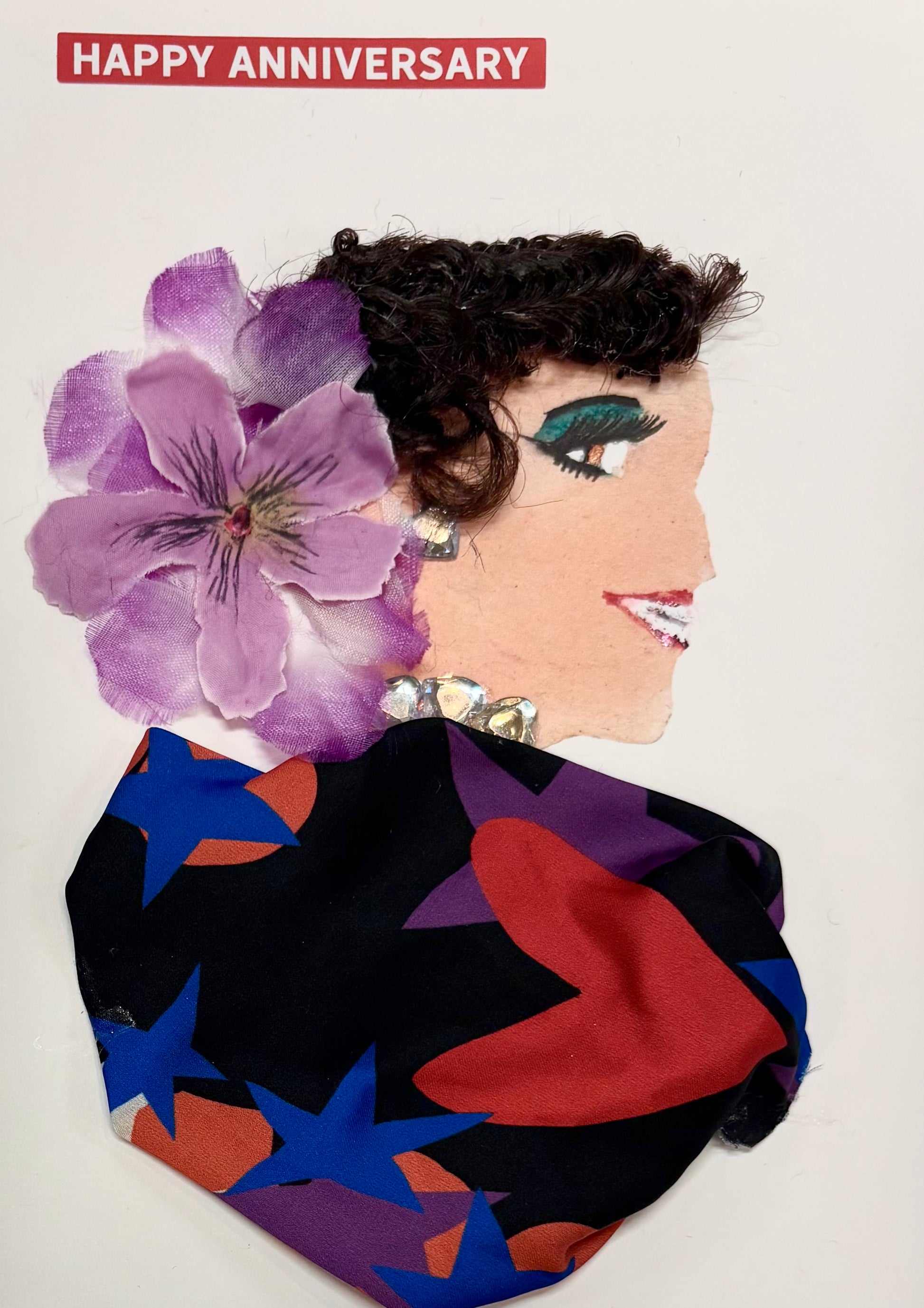 This is a handmade card of a woman named Floral Francesca. She has short, curly hair adorned with a large purple flower. Her earrings and necklace are made of shiny gem-like pieces. She is smiling brightly, with bold green eye makeup and red lips. Her outfit is made from black fabric with a colourful pattern of red hearts, blue stars, and purple accents. At the top, a red and white banner reads "Happy Anniversary."