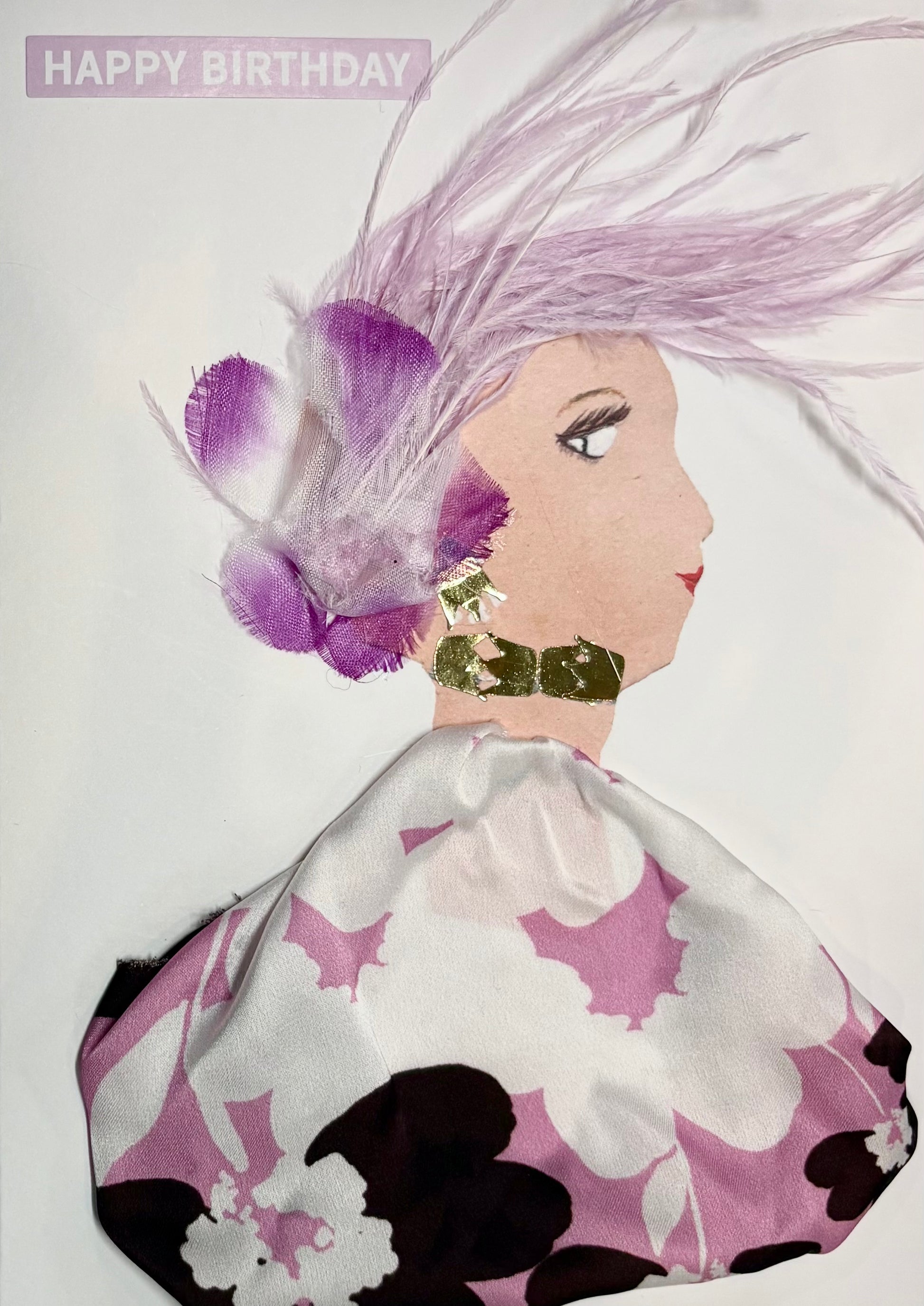 This is a handmade card of a woman named **Fuchsia Feather**. Her flowing lavender feathers sweep to the side, accented with delicate purple and white blossoms. She wears a voluminous dress in a silky white fabric patterned with large floral prints in soft pink, deep plum, and white. Her look is finished with bold, textured gold accessories, and a “Happy Birthday” message appears in the top corner in soft purple.