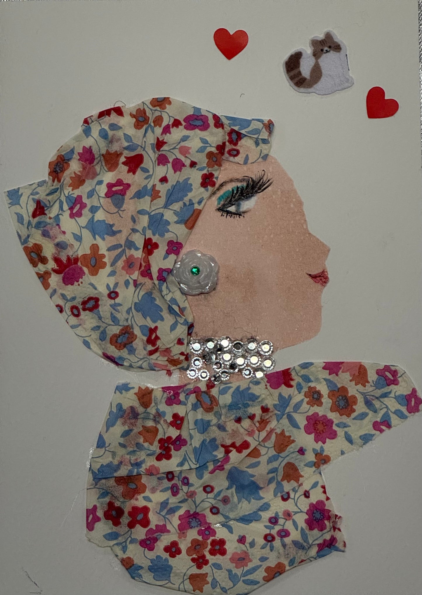 Handmade card that can double as a piece of art featuring a woman wearing a patterned headscarf tied around her hair and a large silver jeweled necklace. The design has a vintage-inspired style with decorative textures and subtle pastel colors. She aslo wears rose shaped earrings that are white with a shiny green bead in the center. On the top right of the card, there is also two red heart designs as well as a small brown and white cat design between the two hearts.