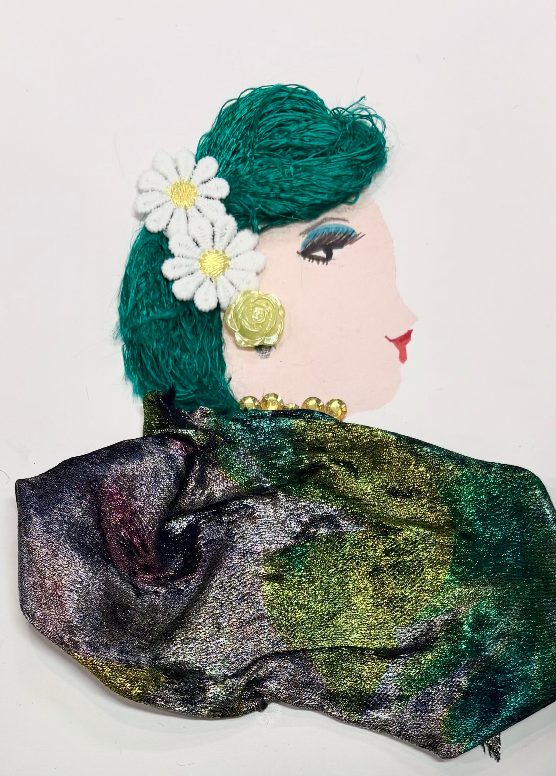 This is a handmade card of a woman named Green Gemma. Her hair is made from green yarn, styled neatly and decorated with two white daisies and a yellow rose. She wears a pearl-like necklace and a shiny, multicoloured fabric in green, gold, and purple wrapped around her shoulders. Her face is drawn in profile with bold eye makeup and red lips.