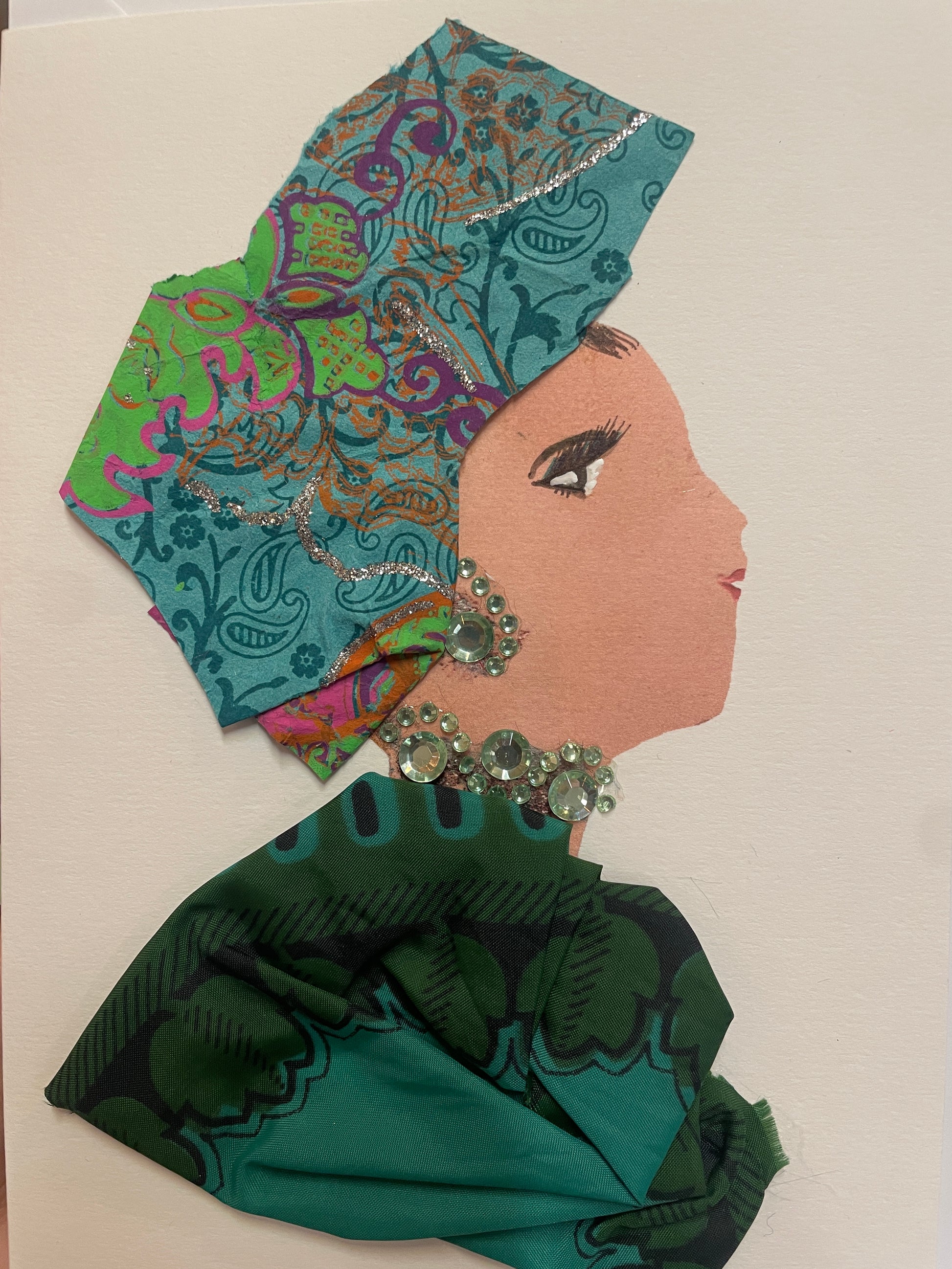 A handmade card showing a woman in profile wearing a richly patterned turquoise and green outfit and matching headwrap. She wears a gem necklace and earring in pale green and silver.