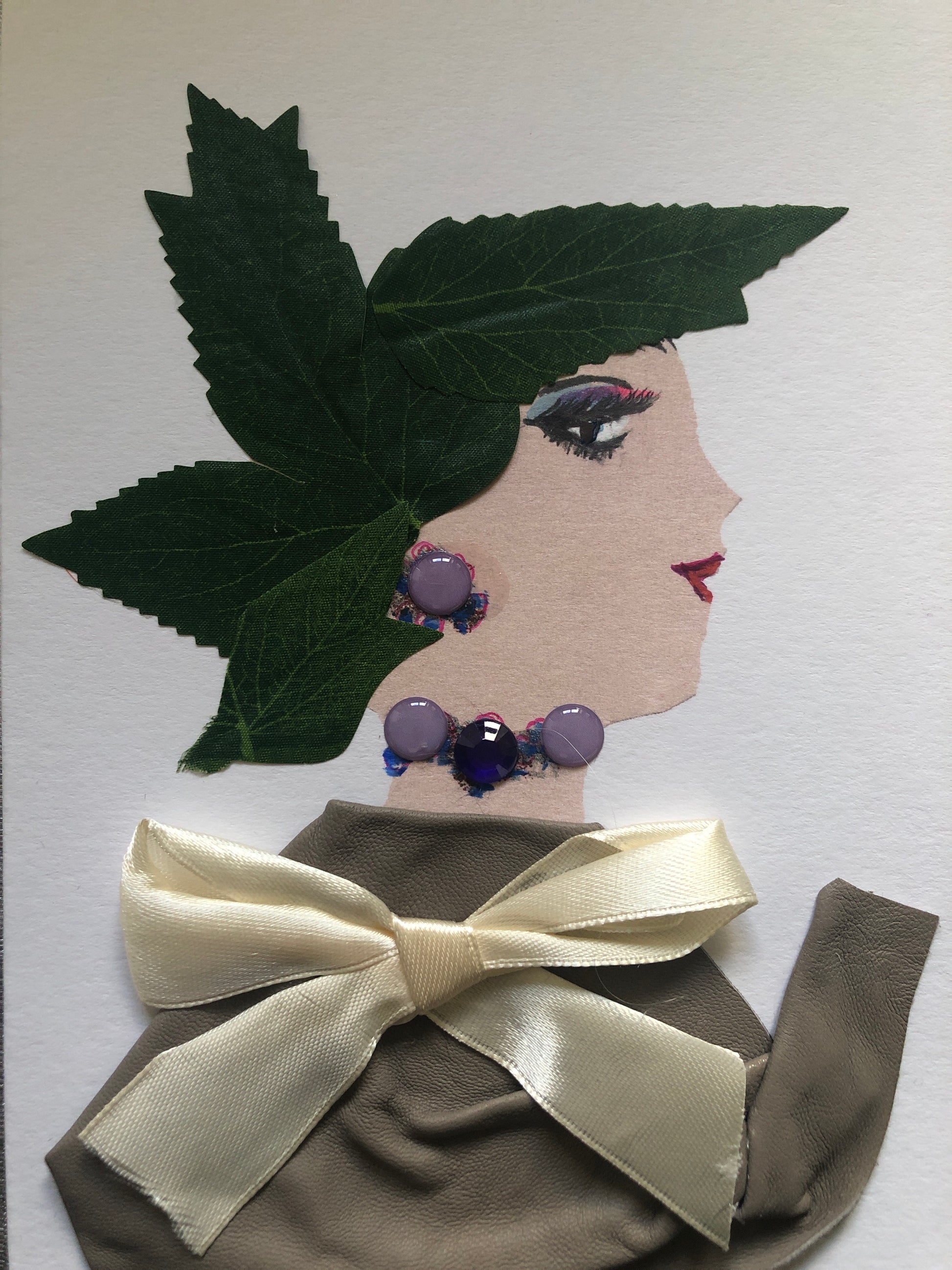 This is a handmade card of a woman named Hampstead Buffet. Her hair is made from green fabric leaves, shaped like a crown. She wears a soft grey-brown outfit tied with a cream-colored satin bow. Her jewelry includes round purple beads on her ear and around her neck.
