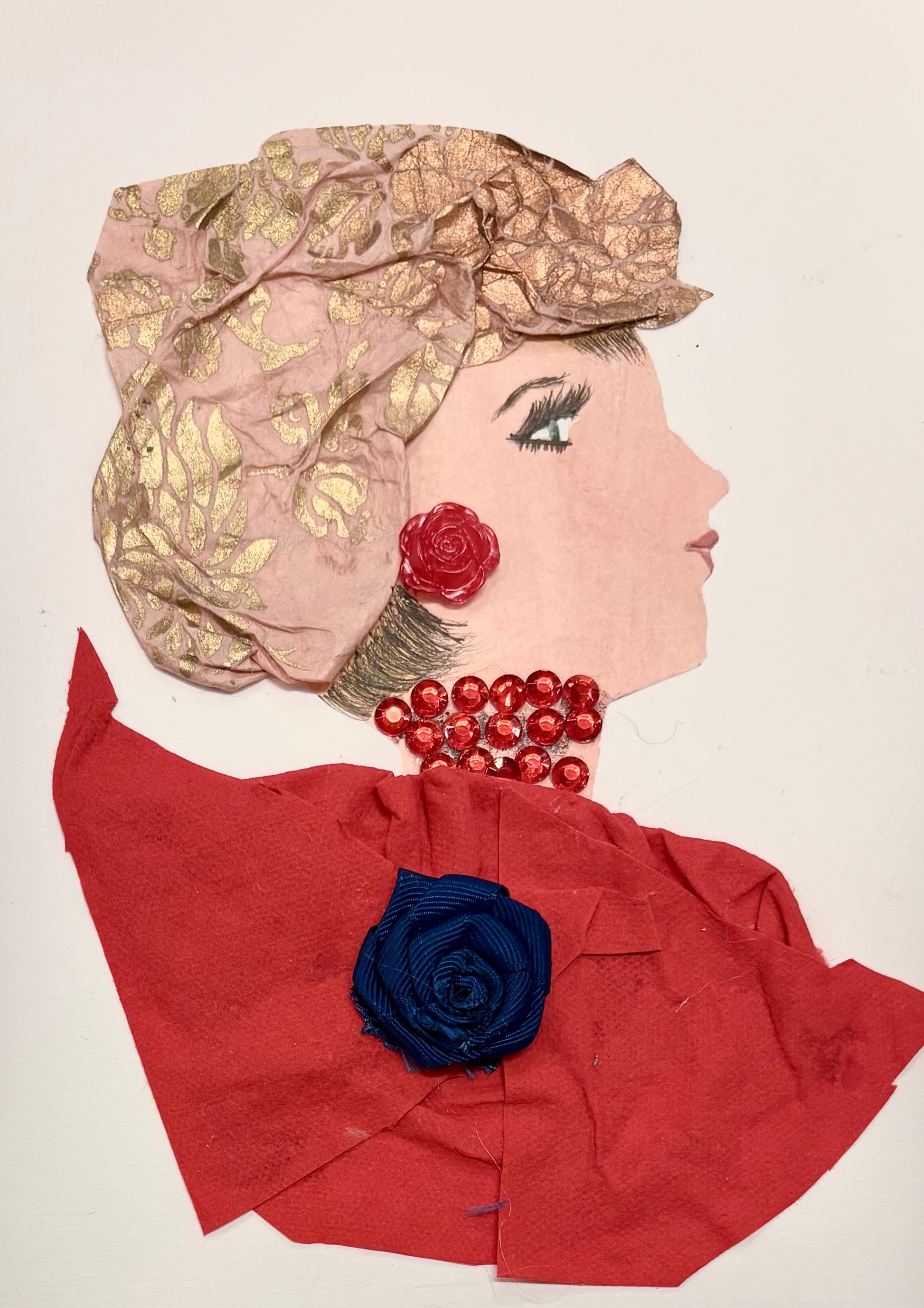 This is a handmade card of a woman named Hannah Hammersmith. She wears a textured pink and gold headwrap with a red rose by her ear. Her necklace is made of shiny red beads, and she is dressed in layered red fabric with a dark blue fabric flower pinned to the front. Her face is shown in profile with neatly drawn features and soft red lips.