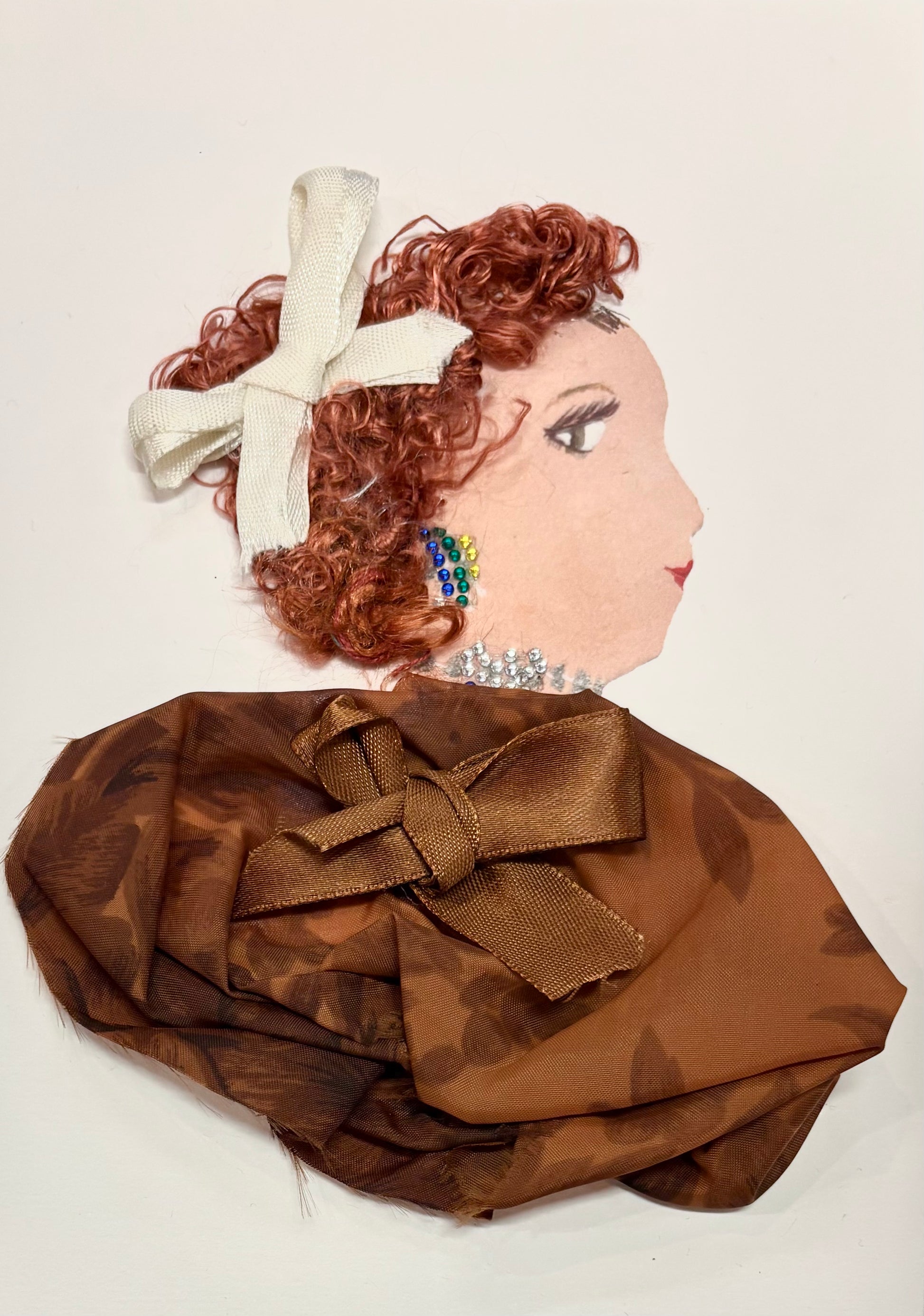 This is a handmade card of a woman named Hazel Hampstead. She has soft reddish-brown curls adorned with a white bow. Her earrings are made of colorful beads—blue, green, and yellow—and she wears a sparkling silver necklace. Her outfit is crafted from brown floral fabric, gathered into gentle folds, and tied with a satin brown ribbon.