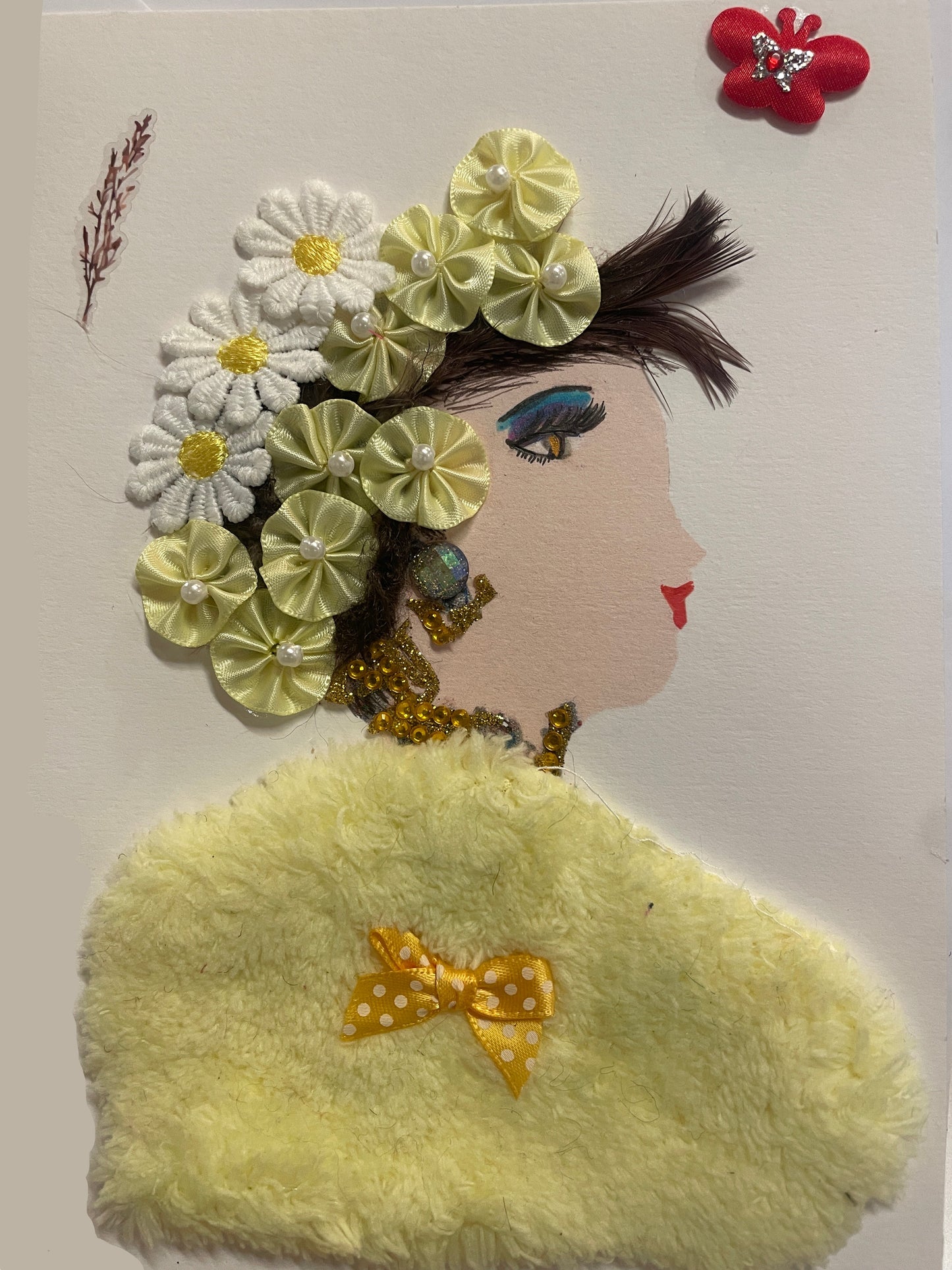 A handmade card features a side-profile portrait of a woman with light skin, red lips, and dramatic blue-and-yellow eye makeup. Her hair is decorated with a crown of fabric flowers in white and pale yellow, each with pearl or yellow button centers. She wears beaded gold jewelry and a large fuzzy yellow shawl with a small polka-dotted yellow bow attached. A red butterfly embellishment and a sprig-like decoration appear near the top of the card.