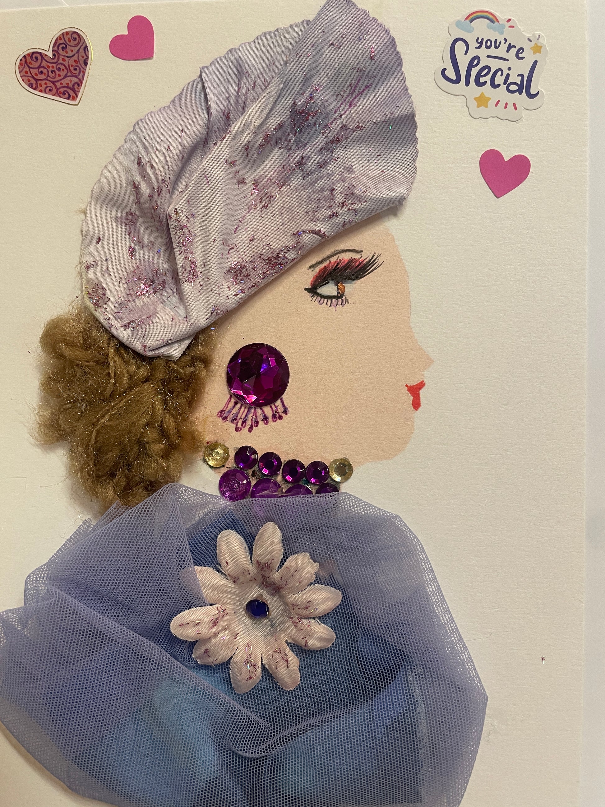 This is a handmade card of a woman named Lilac Lambeth. She wears a glittery lilac hat, a sheer blue wrap with a large flower, and purple gem jewellery. The card is decorated with pink hearts and a sticker that says "You're Special".