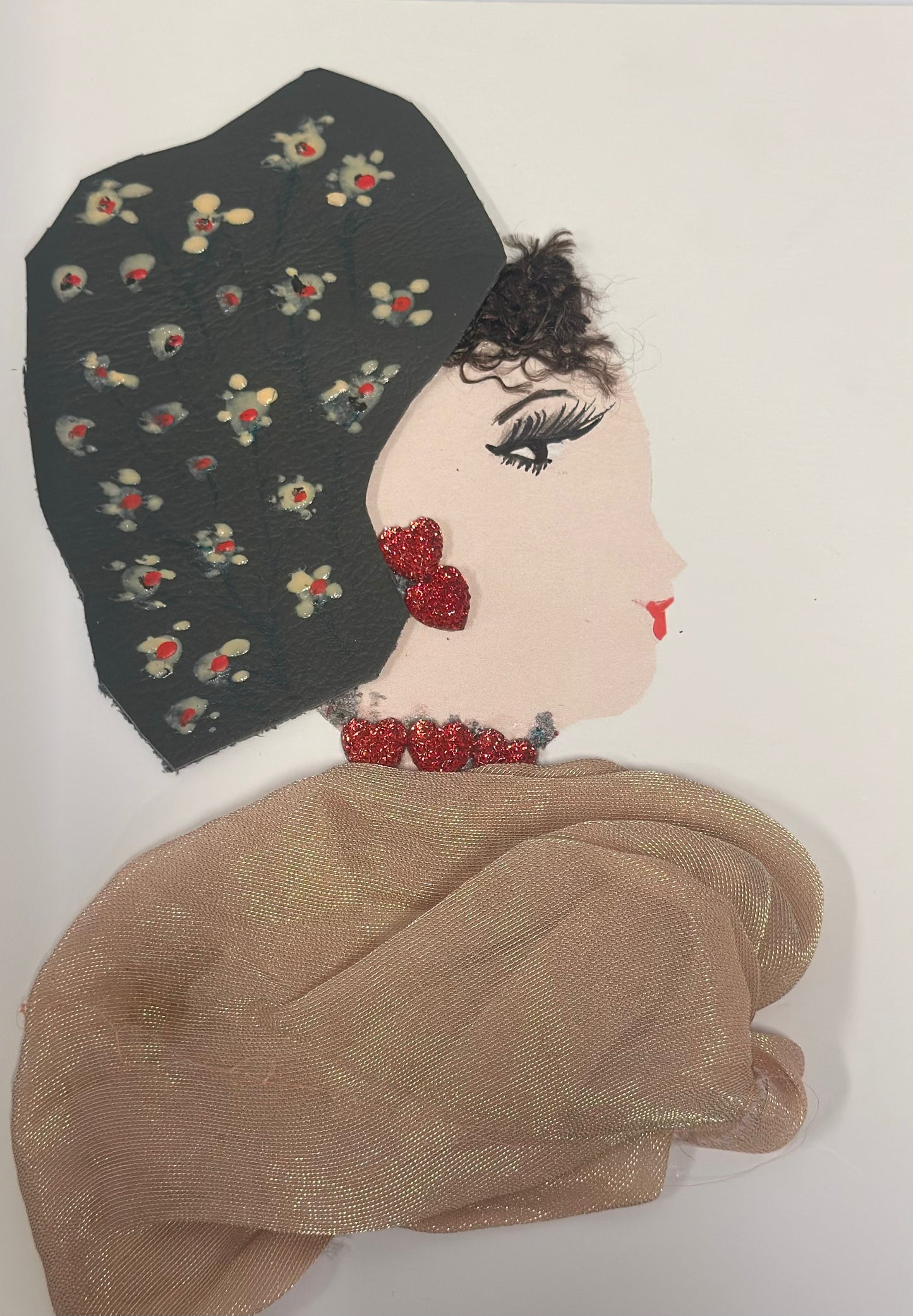 A handmade card features a stylized side-profile portrait of a woman with soft beige skin, long black eyelashes, and red lips. She wears a black headscarf decorated with small cream and red floral patterns, glittery red heart-shaped earrings, and a matching red beaded choker. Her textured brown hair peeks out from under the scarf, and she is draped in a shimmery beige fabric shawl that adds depth and texture to the design