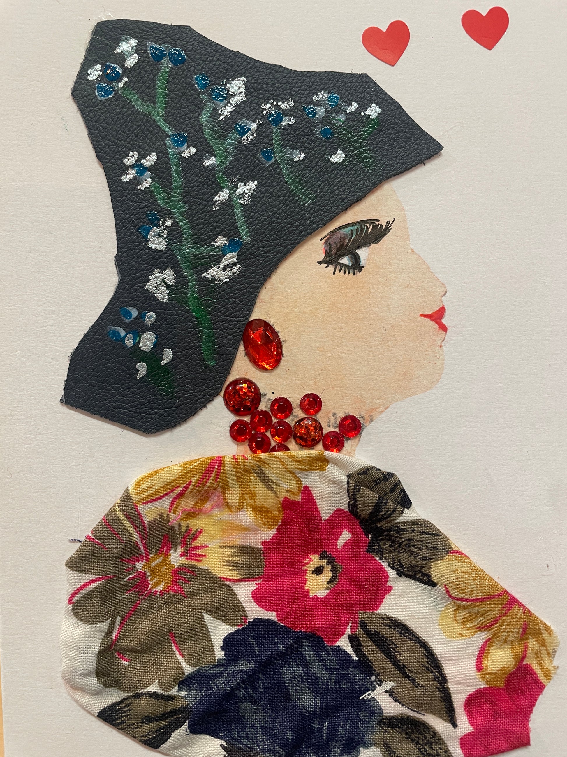 This is a handmade card of a woman named Amira Abbeygate. She wears a black hat with hand-painted blue and white flowers, a floral-print blouse, and large red gem jewellery. There are two red heart stickers in the top right corner.