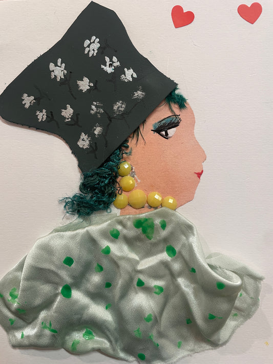 This is a handmade card of a woman named Saffron Southwark. She wears a grey hat with flower paintings, a light green blouse with green splatters, large yellow gem jewellery, and has blue-green hair peeking out. Two red heart stickers are in the top right corner.