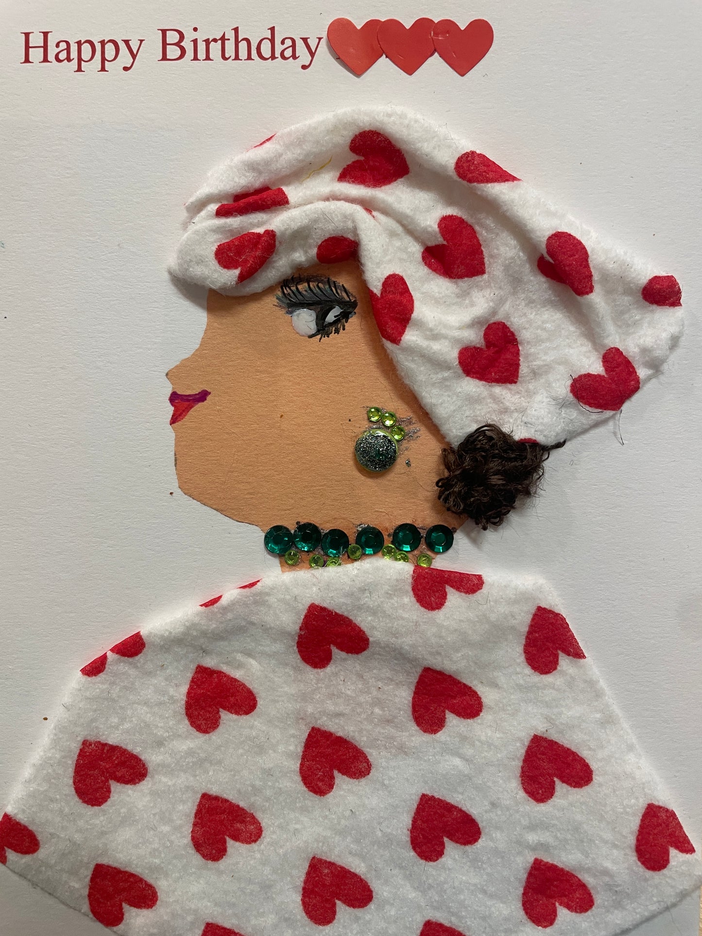 This is a handmade card of a woman named Nia Northcote. She wears a white hat and blouse with red hearts printed on them, green gem jewellery, and a "Happy Birthday" sticker in the top left corner with three red hearts around it.