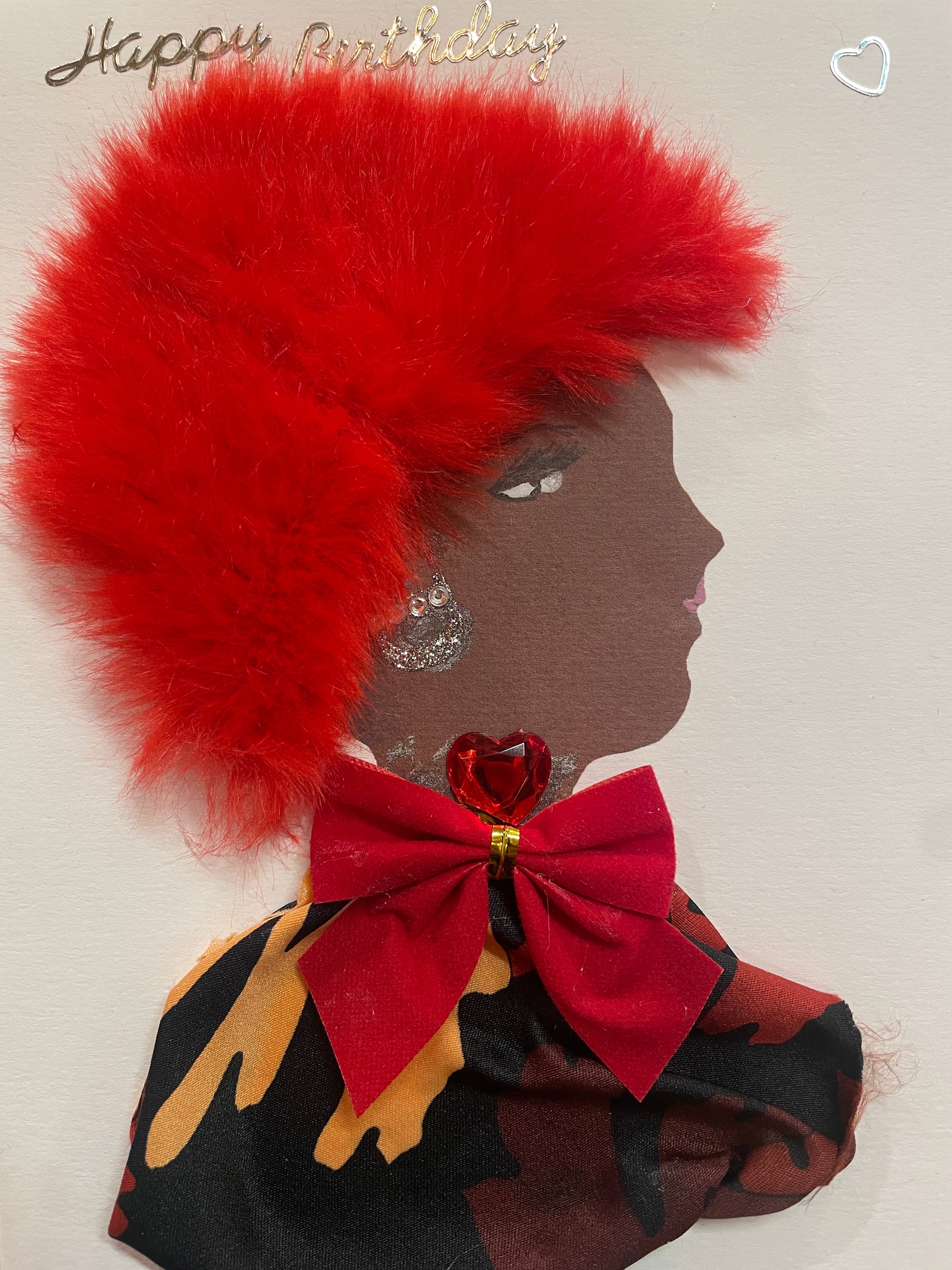 This is a handmade card of a woman named Ruby Rotherhithe. She has big red hair, a patterned blouse with a large red bow, red gem jewellery, a “Happy Birthday” sticker in one corner, and a heart sticker in the other.