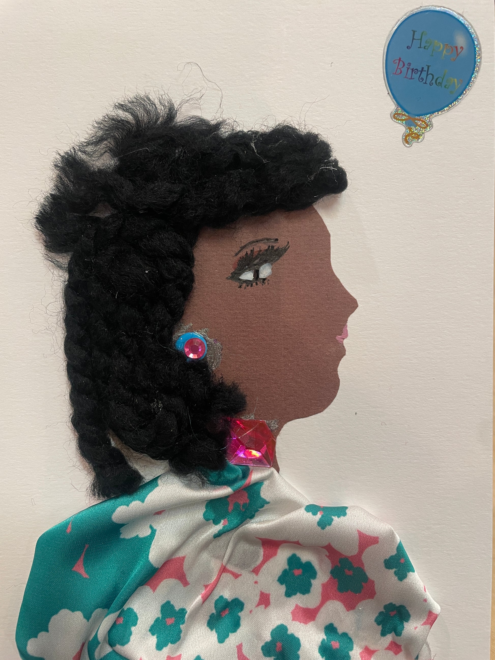This is a handmade card of a woman named Dahlia Deptford. She wears a blue blouse with colourful flowers, pink gem jewellery, black hair, and a “Happy Birthday” balloon sticker in the corner.
