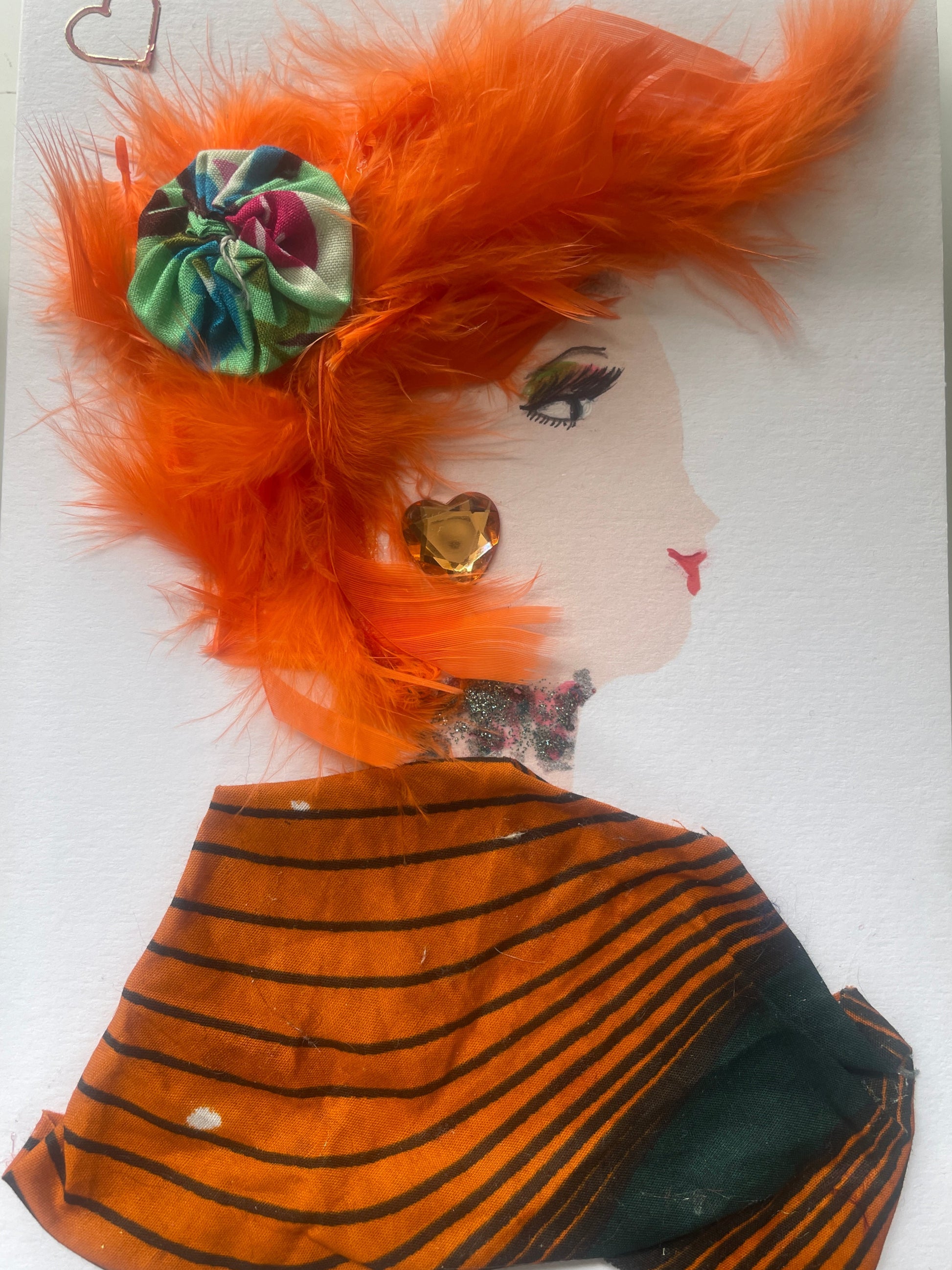 This is a handmade card of a woman named AmberLe Archway. She has bright orange feathered hair with a colourful rosette, a striped orange and black wrap, yellow gem jewellery, and a small heart sticker.